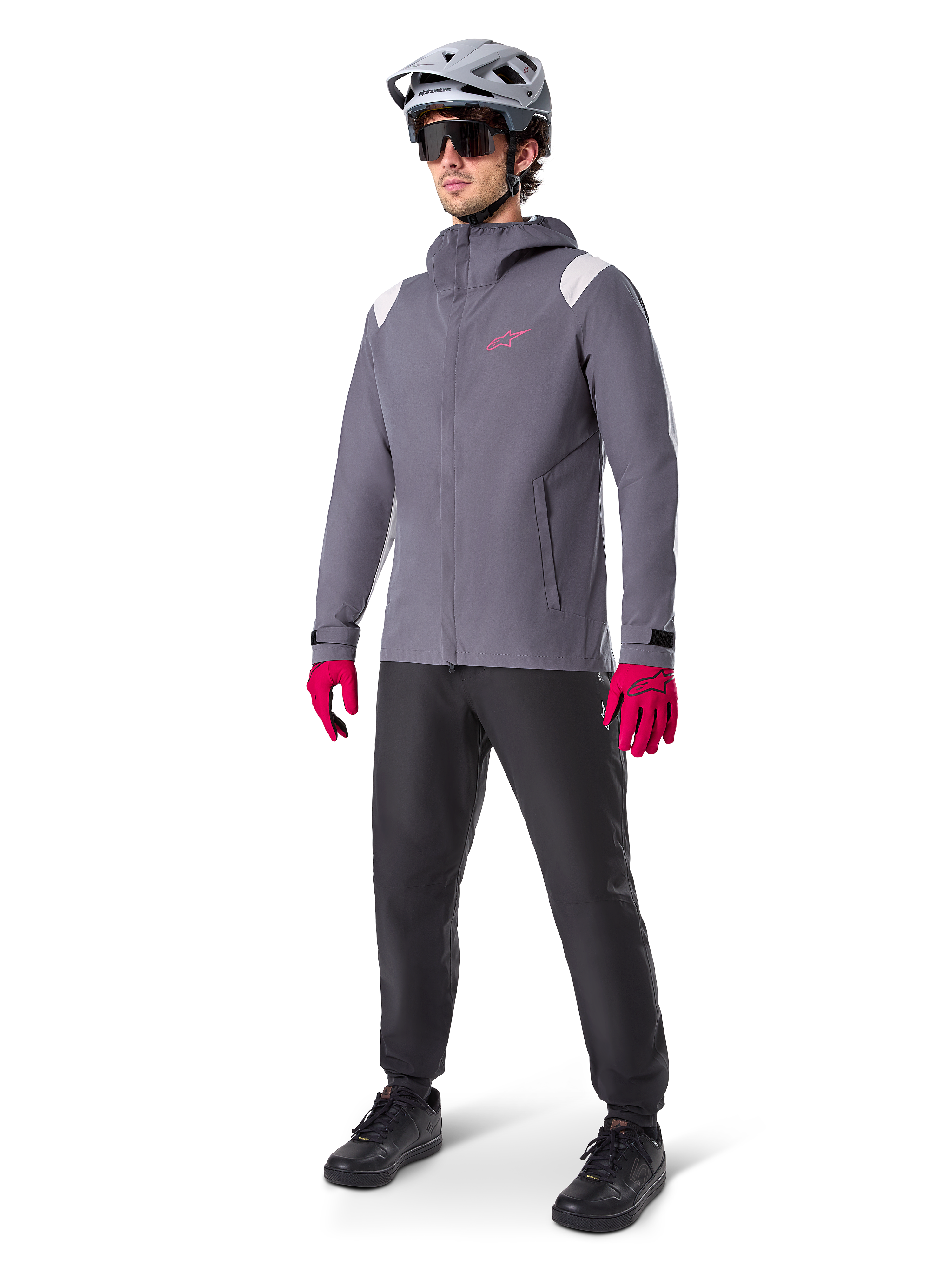 Modelo masculino con la chaqueta impermeable Alpinestars A-Dura en color Dark Titanium, chaqueta gris con capucha, logotipo rosa en el pecho y paneles gris claro en los hombros, combinada con casco de MTB gris, gafas de sol oscuras, guantes rosa vibrante y pantalones negros para ciclismo de montaña.