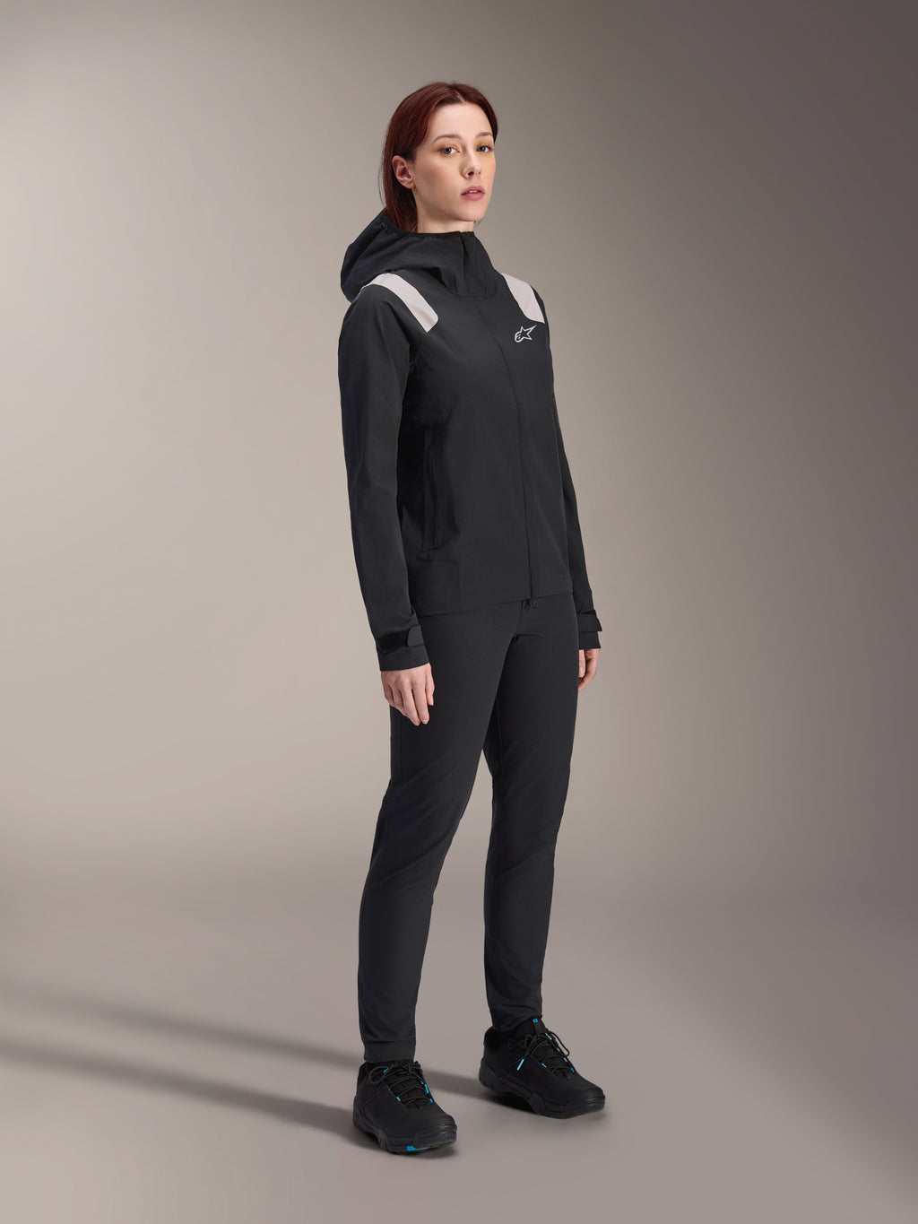 Mujer vistiendo la chaqueta de lluvia Alpinestars Stella A-Dura en color negro, capa exterior impermeable de 2.5 capas para MTB con detalles en gris claro en los hombros, pantalones negros y zapatillas de ciclismo a juego, retrato de estudio para equipamiento de ciclismo de alto rendimiento.