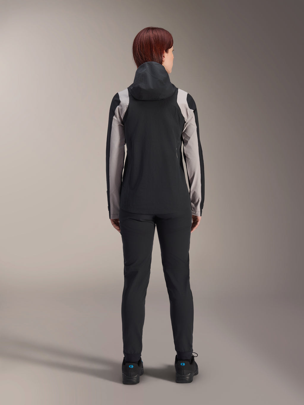 Ciclista femenina vista desde atrás con la chaqueta de lluvia Alpinestars Stella A-Dura, negra con paneles gris claro en las mangas y los hombros, con la capucha puesta, combinada con pantalones negros y zapatillas de ciclismo, equipamiento técnico de lluvia para MTB