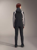 Ciclista femenina vista desde atrás con la chaqueta de lluvia Alpinestars Stella A-Dura, negra con paneles gris claro en las mangas y los hombros, con la capucha puesta, combinada con pantalones negros y zapatillas de ciclismo, equipamiento técnico de lluvia para MTB