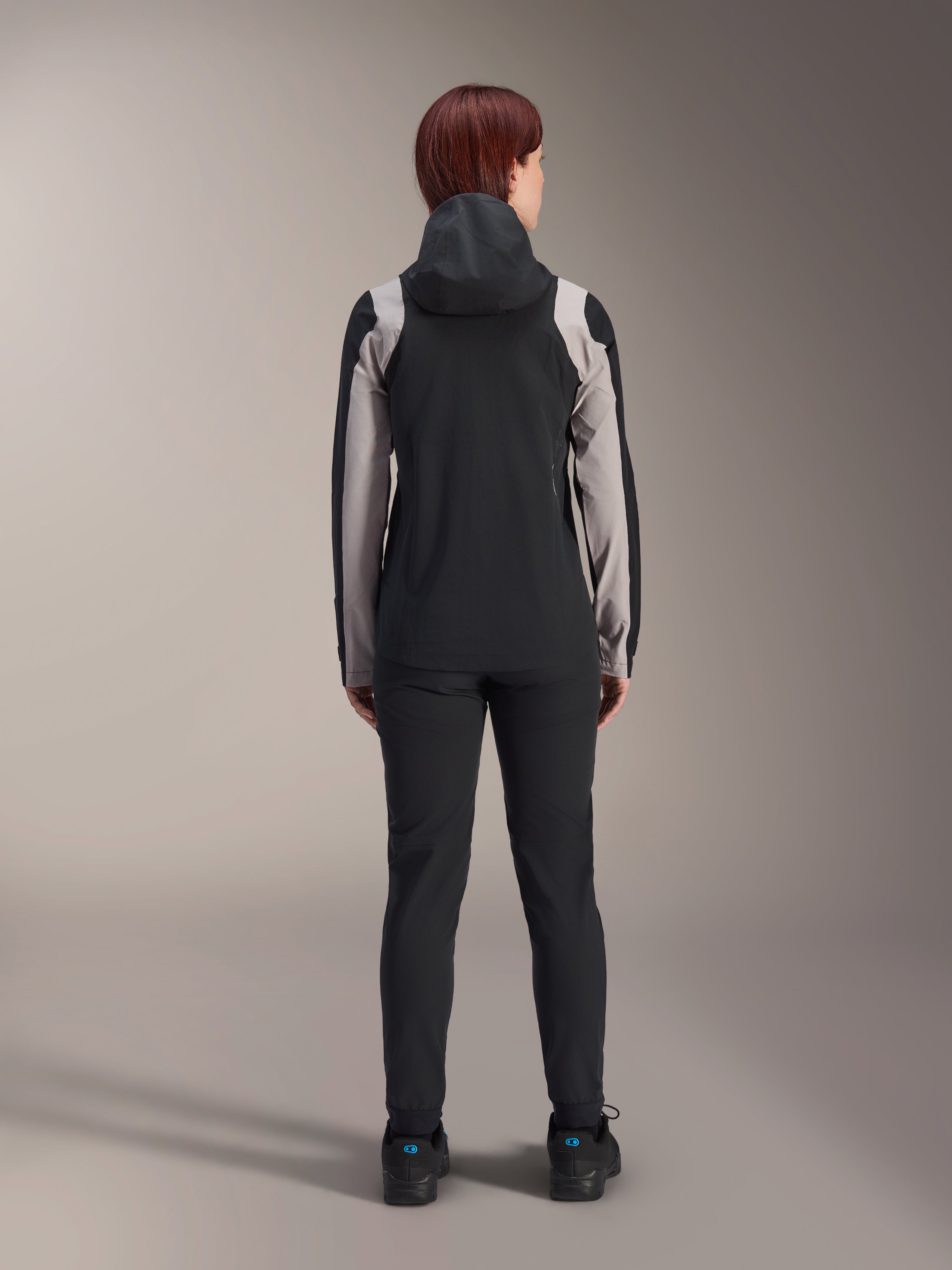 Ciclista femenina vista desde atrás con la chaqueta de lluvia Alpinestars Stella A-Dura, negra con paneles gris claro en las mangas y los hombros, con la capucha puesta, combinada con pantalones negros y zapatillas de ciclismo, equipamiento técnico de lluvia para MTB