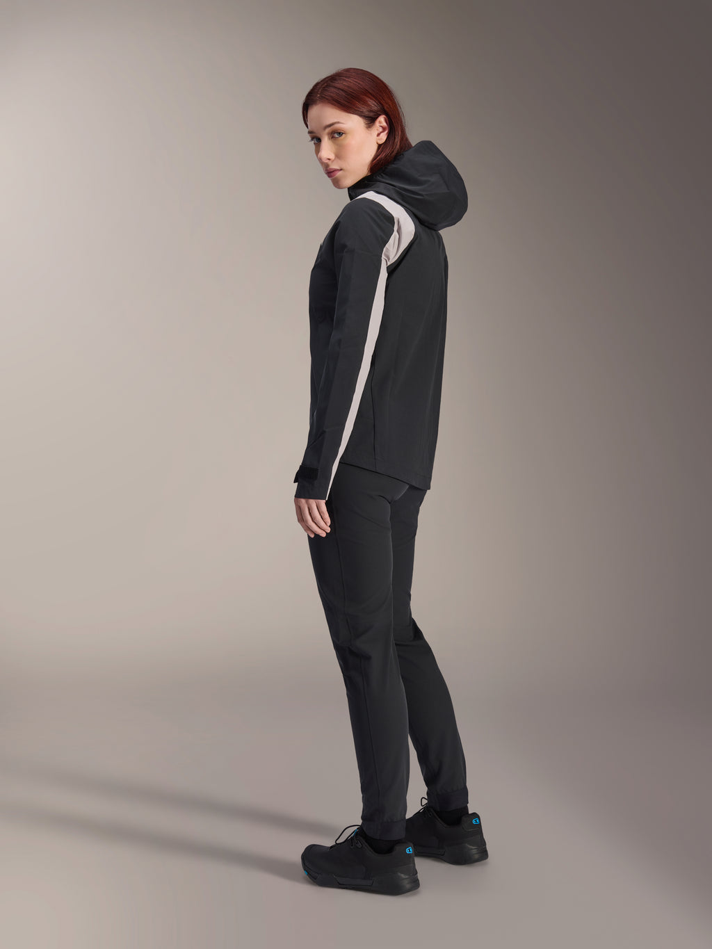 Mujer con chaqueta de lluvia Alpinestars Stella A-Dura, color negro con franja gris claro en la manga, chaqueta de ciclismo impermeable con capucha para MTB, combinada con pantalones técnicos negros y calzado de ciclismo negro, vista de perfil en estudio
