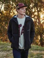 Gorra Trucker Backline de Alpinestars en colores teja, negro y mostaza, llevada por un hombre con una chaqueta negra con capucha y camiseta con gráfico, estilo urbano casual inspirado en el motociclismo en un entorno exterior durante la hora dorada