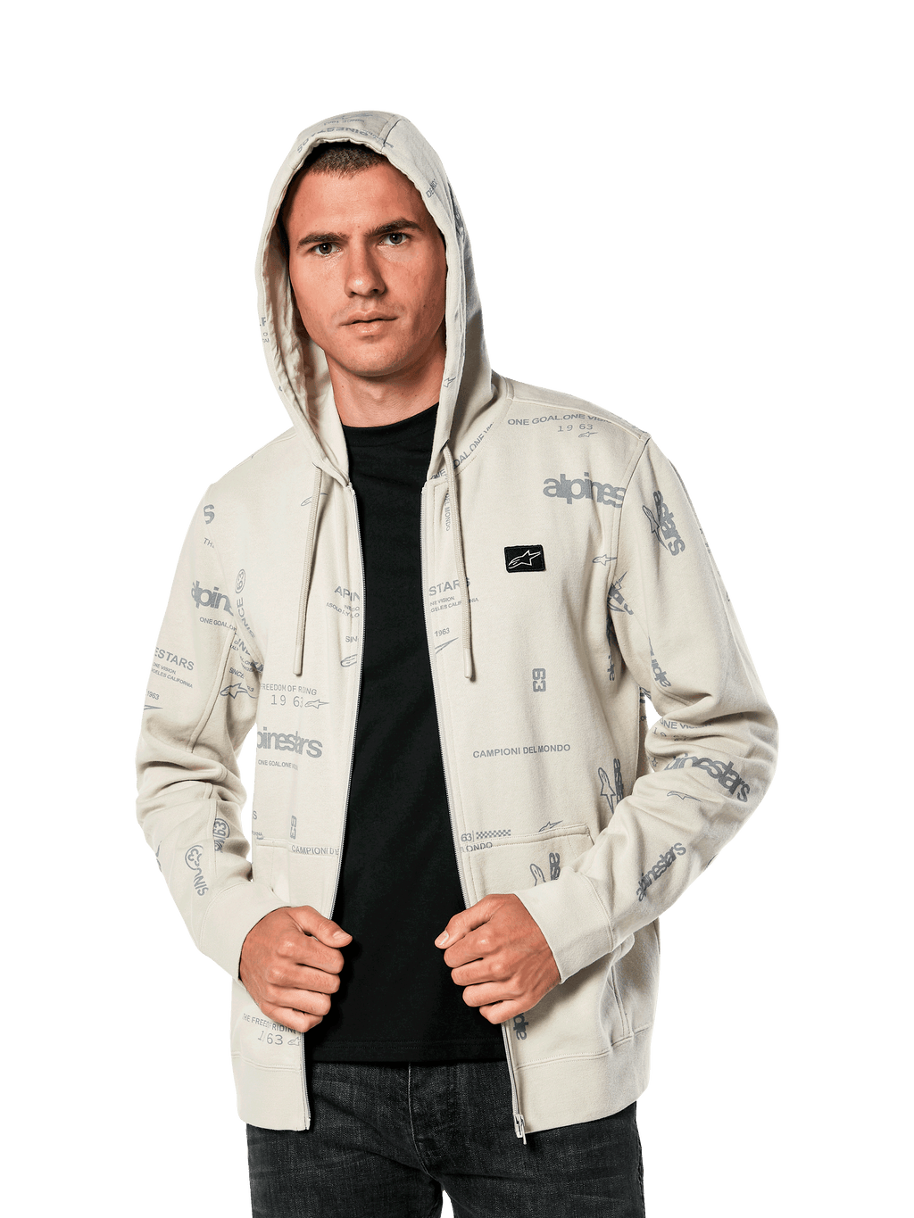 Sudadera con capucha Plenitude de Alpinestars, ropa deportiva casual, sudadera con cremallera frontal en blanco roto con gráficos de logotipos grises por toda la prenda, usada por un hombre con la capucha puesta sobre una camiseta negra, con un pequeño parche con el logotipo en el pecho y bolsillo tipo canguro