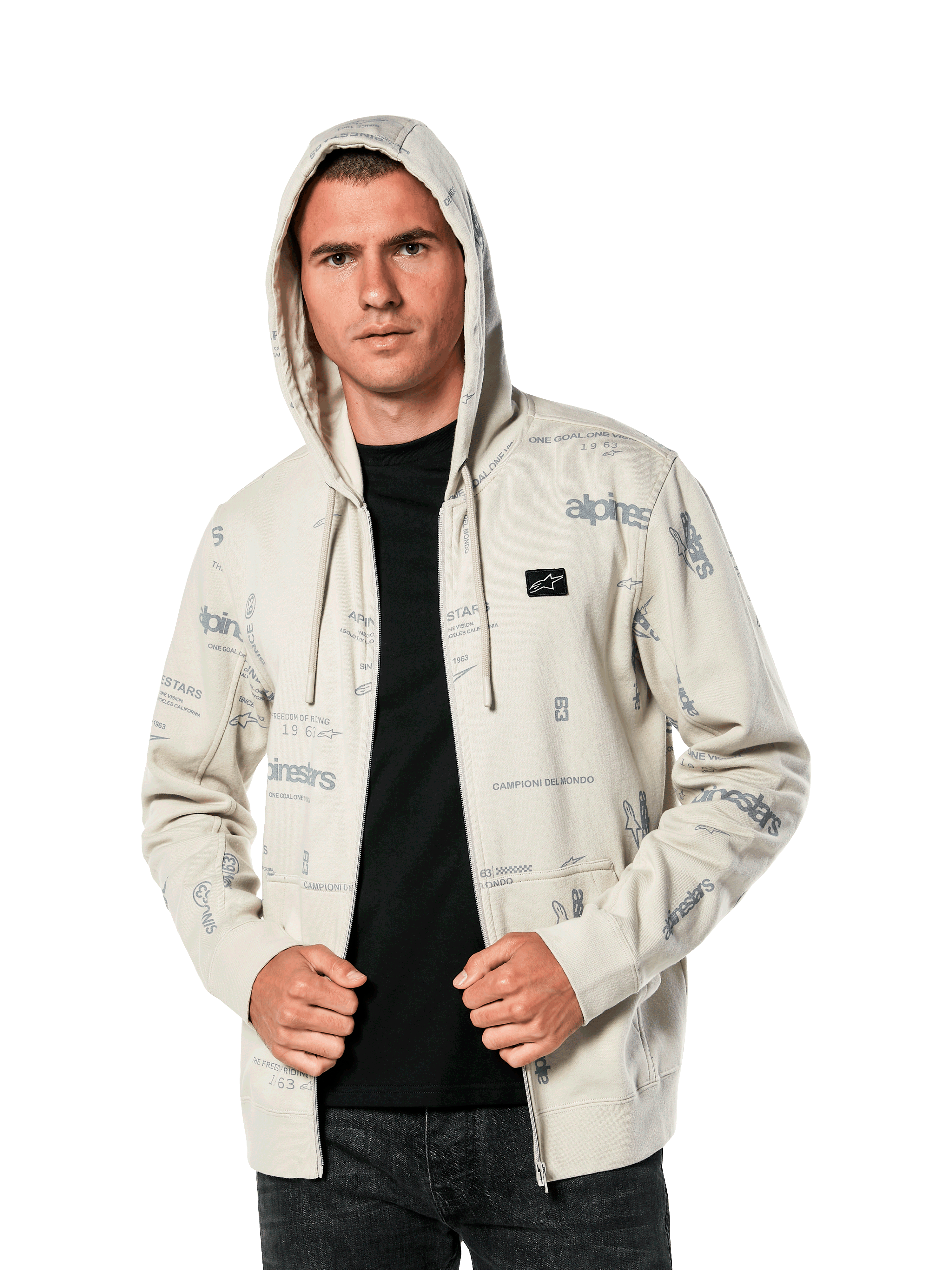 Sudadera con capucha Plenitude de Alpinestars, ropa deportiva casual, sudadera con cremallera frontal en blanco roto con gráficos de logotipos grises por toda la prenda, usada por un hombre con la capucha puesta sobre una camiseta negra, con un pequeño parche con el logotipo en el pecho y bolsillo tipo canguro
