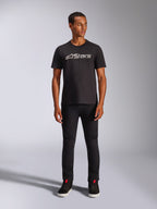 Camiseta Alpinestars Blaze 2.0 CSF, camiseta de manga corta, negra con logotipo frontal en gris y blanco, lucida por un modelo con pantalones negros y calzado negro, ropa urbana informal inspirada en el automovilismo