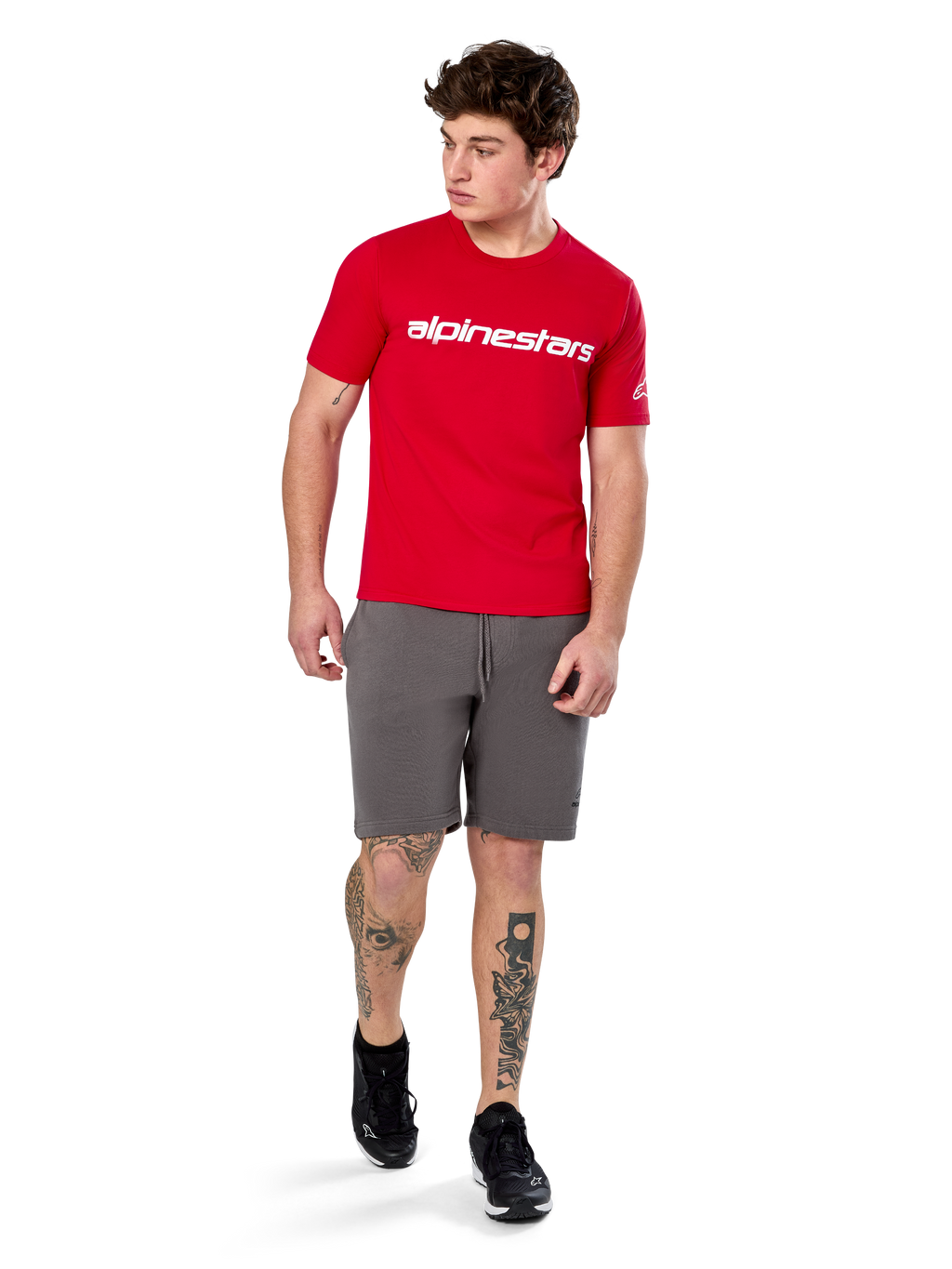 Camiseta Alpinestars Linear Wordmark 2.0 CSF, camiseta de manga corta, roja con logotipo de marca lineal blanco en el pecho, usada por un modelo con pantalones cortos grises y zapatillas negras, ropa urbana casual inspirada en el automovilismo