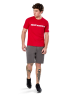 Camiseta Alpinestars Linear Wordmark 2.0 CSF, camiseta de manga corta, roja con logotipo de marca lineal blanco en el pecho, usada por un modelo con pantalones cortos grises y zapatillas negras, ropa urbana casual inspirada en el automovilismo