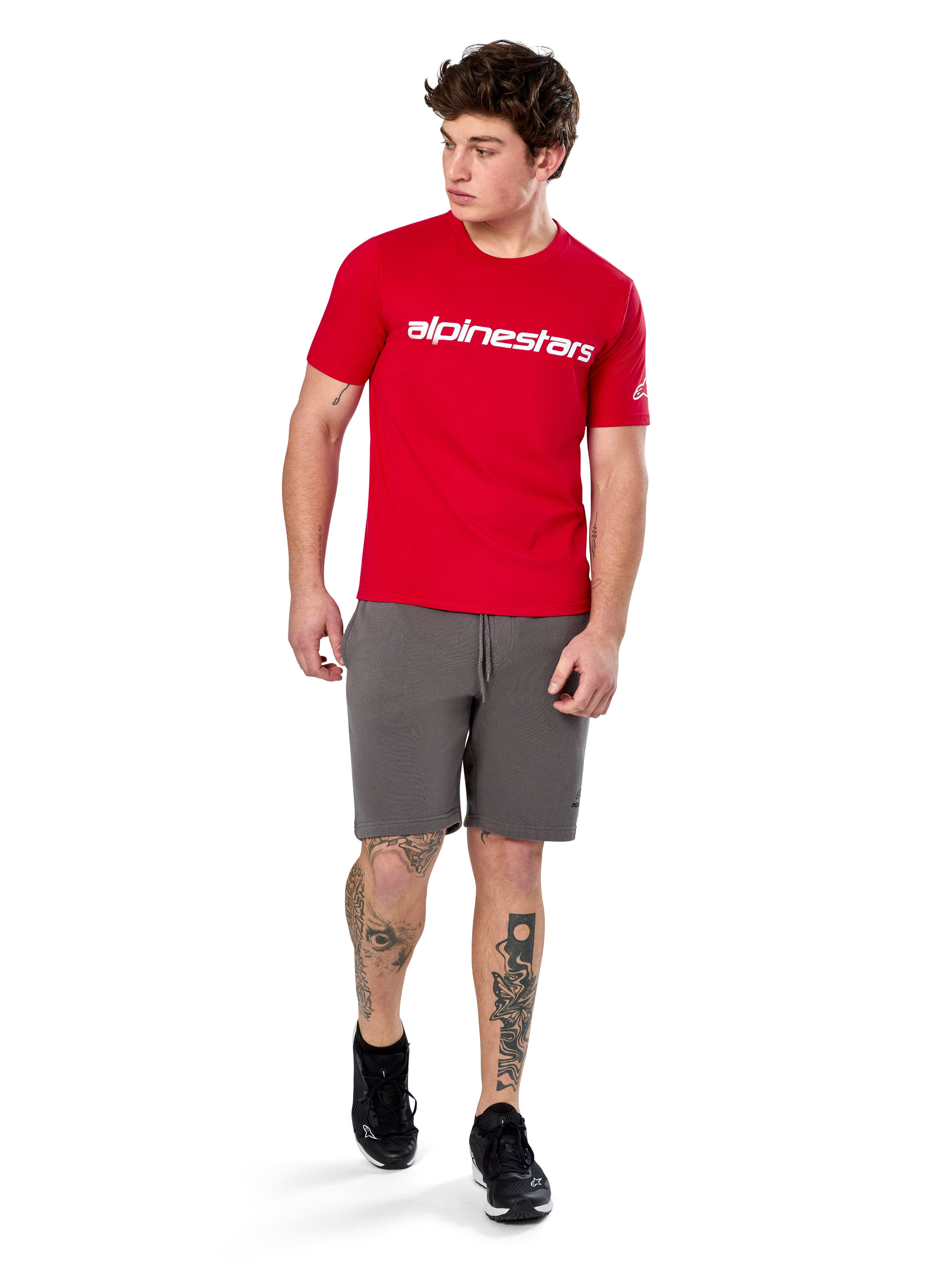 Camiseta Alpinestars Linear Wordmark 2.0 CSF, camiseta de manga corta, roja con logotipo de marca lineal blanco en el pecho, usada por un modelo con pantalones cortos grises y zapatillas negras, ropa urbana casual inspirada en el automovilismo
