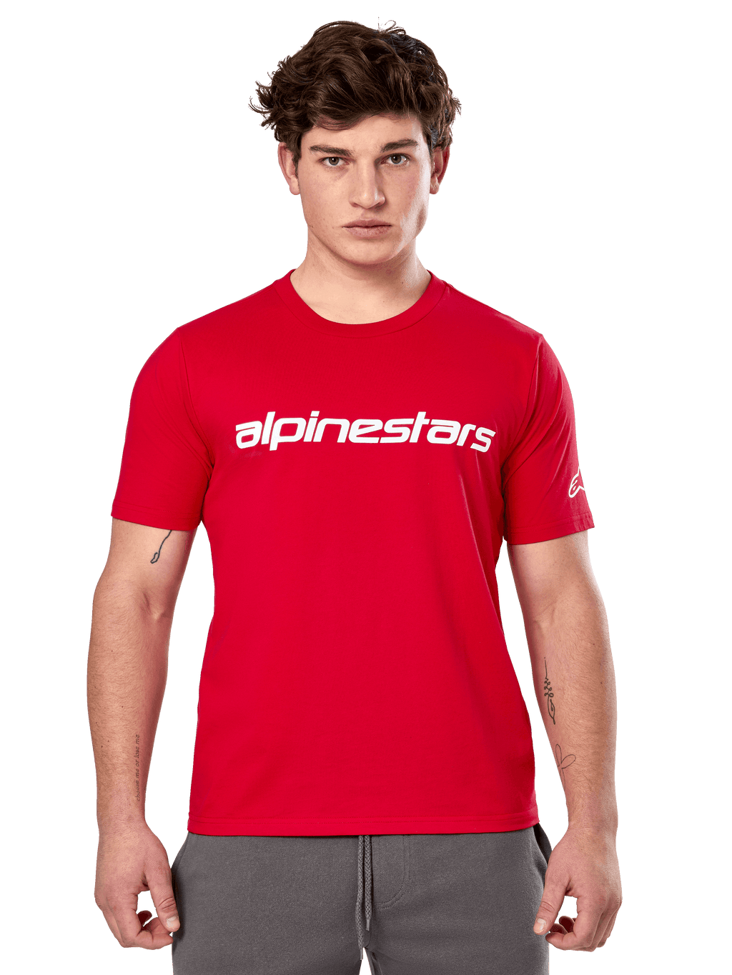 Alpinestars Linear Wordmark 2.0 CSF Tee, camiseta de manga corta roja, marca de palabra lineal blanca en el pecho y logotipo Ageless en la manga, lucida por un modelo masculino con pantalones de chándal grises, estilo urbano casual inspirado en el automovilismo