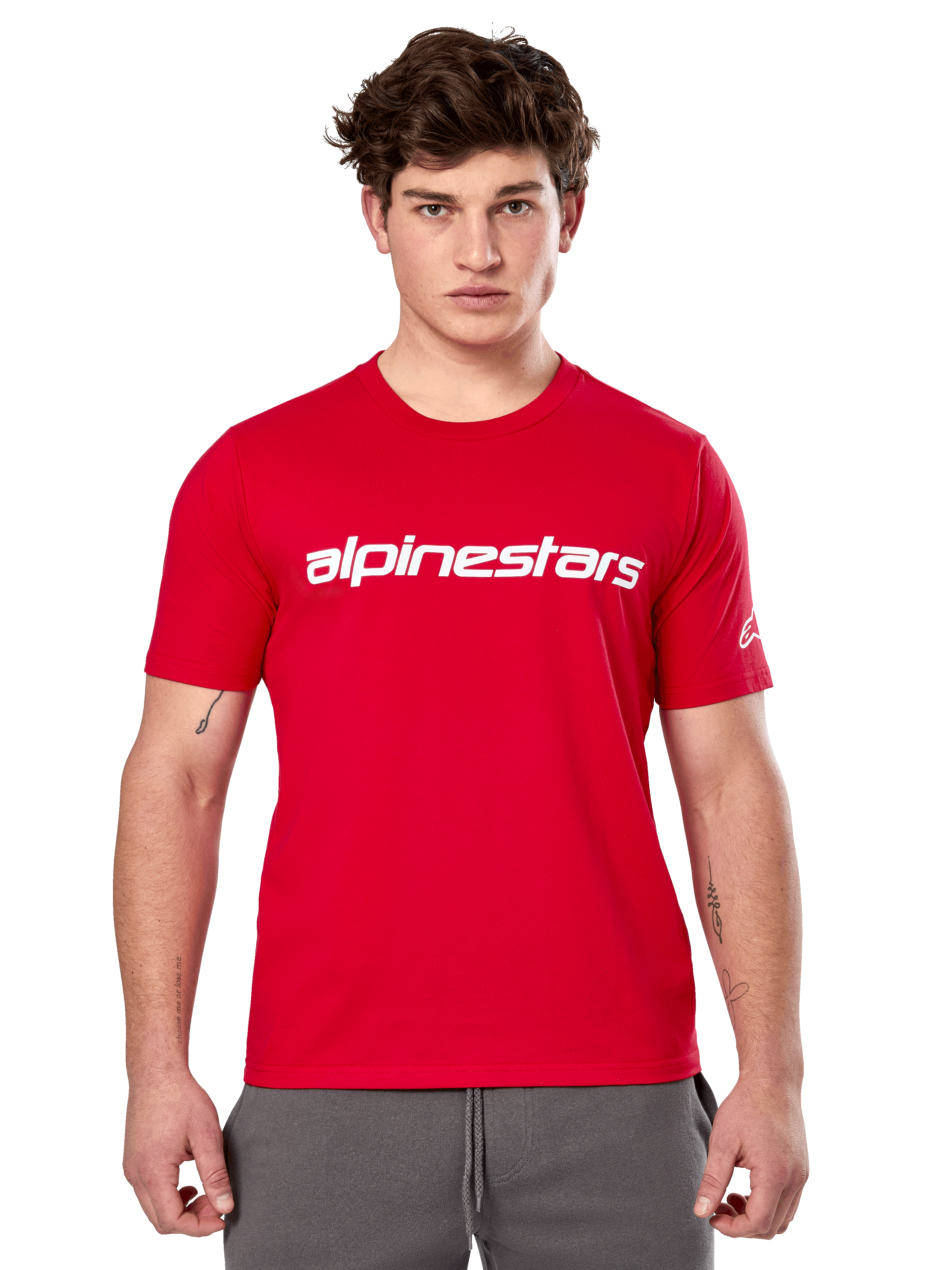 Alpinestars Linear Wordmark 2.0 CSF Tee, camiseta de manga corta roja, marca de palabra lineal blanca en el pecho y logotipo Ageless en la manga, lucida por un modelo masculino con pantalones de chándal grises, estilo urbano casual inspirado en el automovilismo