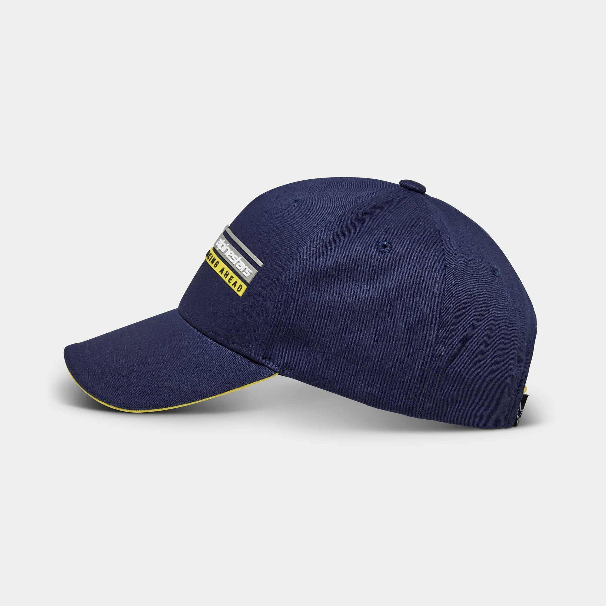Gorra Alpinestars Par Hat, gorra de béisbol azul marino, vista lateral, corona estructurada con visera curva y detalle sándwich amarillo, bordado 3D del logotipo lineal Racing Ahead, accesorio casual inspirado en el automovilismo
