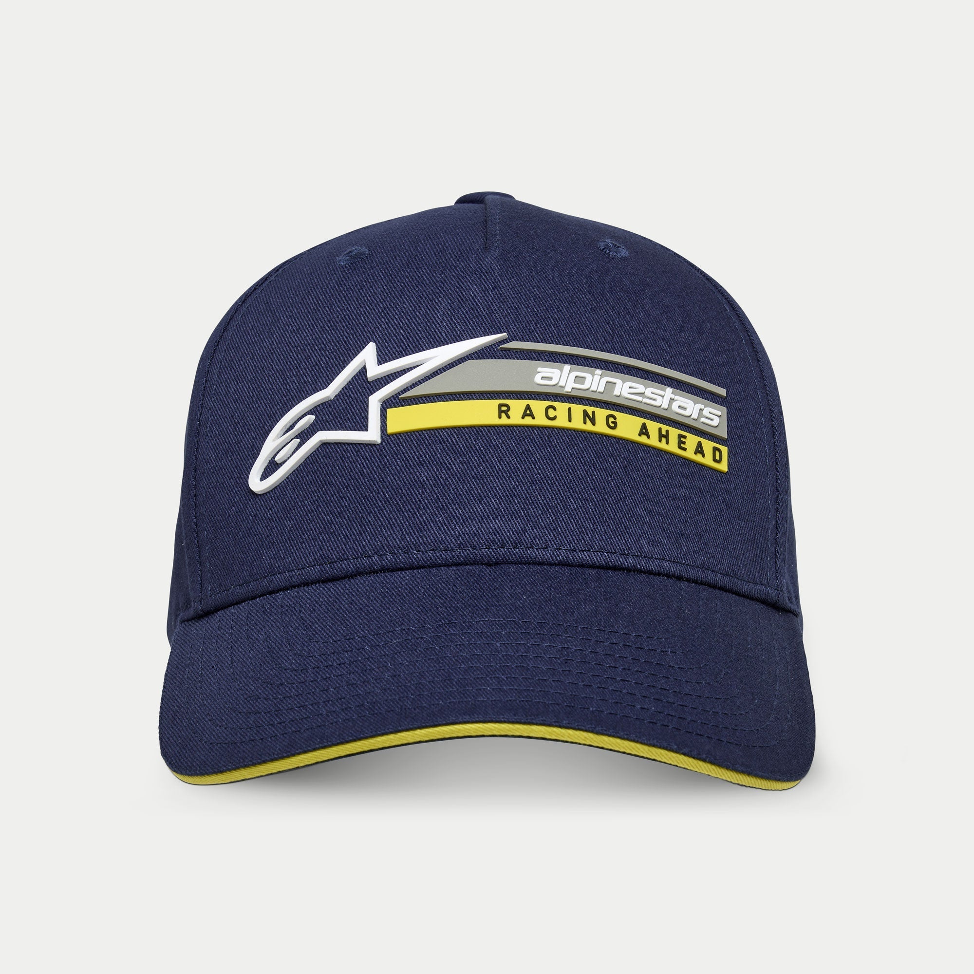 Gorra Par de Alpinestars, azul marino, gorra estructurada tipo snapback con visera curva, con logotipo Astars 3D blanco y gráfico 'alpinestars RACING AHEAD' en gris y amarillo, detalle de visera tipo sándwich amarilla, estilo urbano casual inspirado en el motor