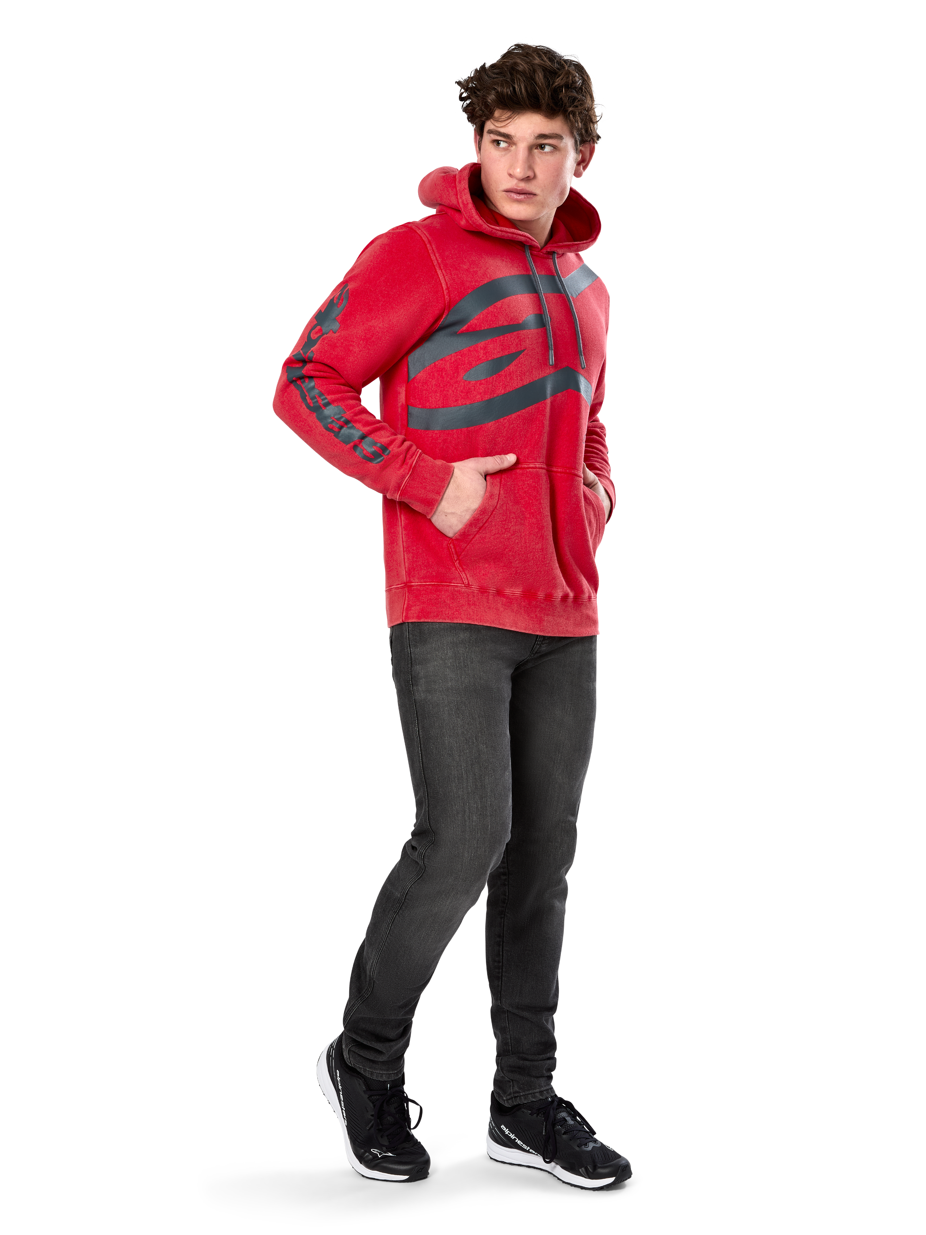 Sudadera con capucha Alpinestars Unbound, forro polar rojo teñido en prenda, logotipo oscuro extragrande en el pecho y marca en las mangas, bolsillo canguro, capucha con cordón, usada por un modelo con vaqueros gris oscuro y zapatillas negras, ropa urbana casual inspirada en el automovilismo