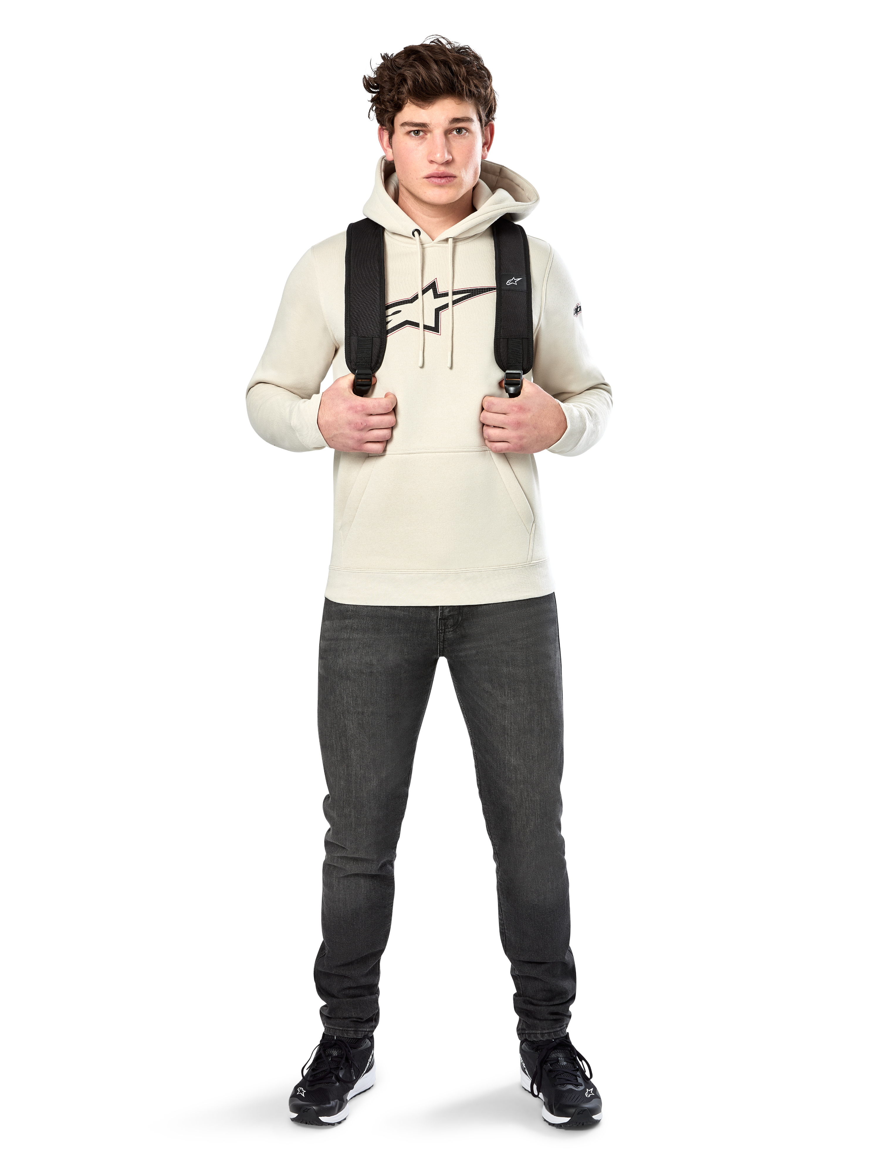 Sudadera con capucha Alpinestars Insignia, ropa urbana casual, sudadera de forro polar Off White, logotipo con apariencia de fibra de carbono y contorno rojo, capucha con cordón, bolsillo canguro, vestida por un modelo con mochila negra y vaqueros gris oscuro