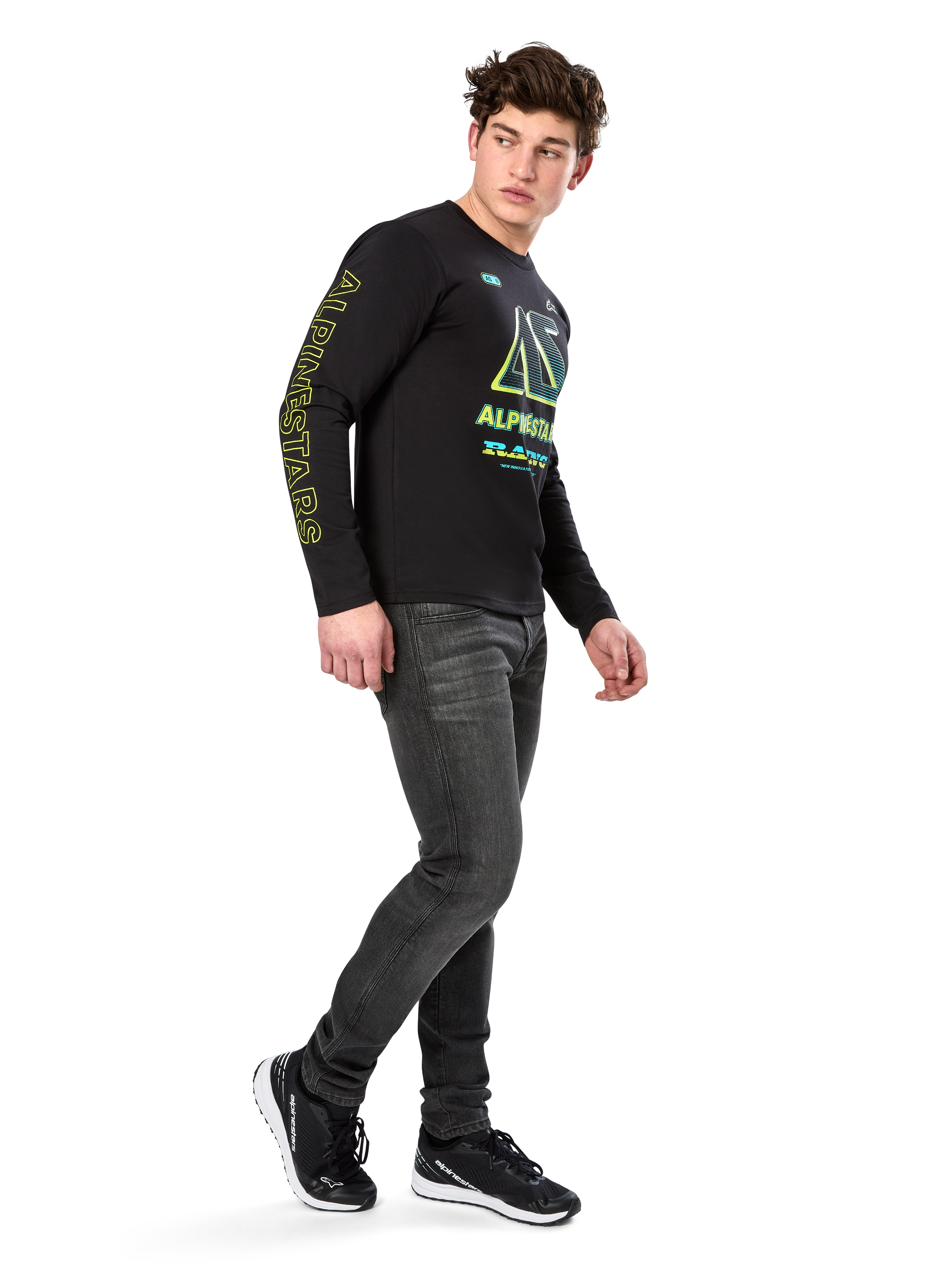 Man wearing Alpinestars Ayess CSF Long Sleeve Tee, Black, featuring yellow sleeve lettering and vibrant lime and cyan chest graphics, paired with dark grey jeans and black sneakers, casual motorsport-inspired streetwear.