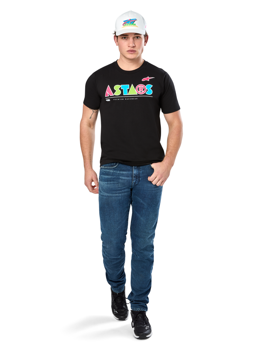 Modelo masculino vistiendo la camiseta Alpinestars Cereal CSF, camiseta negra de manga corta con un colorido logo geométrico de ASTARS, combinada con pantalones vaqueros azules y gorra blanca, estilo streetwear casual inspirado en el automovilismo.