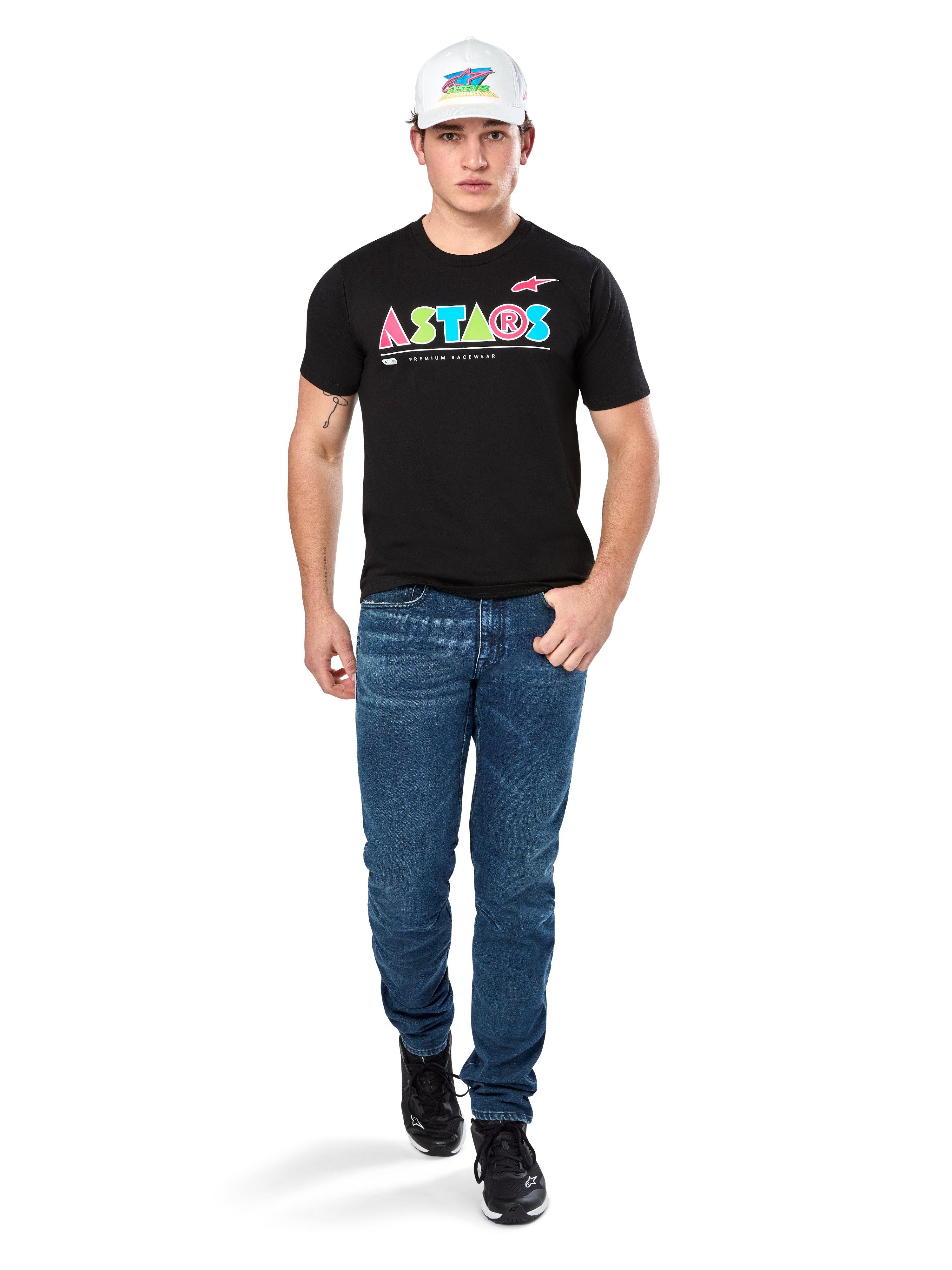 Modelo masculino vistiendo la camiseta Alpinestars Cereal CSF, camiseta negra de manga corta con un colorido logo geométrico de ASTARS, combinada con pantalones vaqueros azules y gorra blanca, estilo streetwear casual inspirado en el automovilismo.