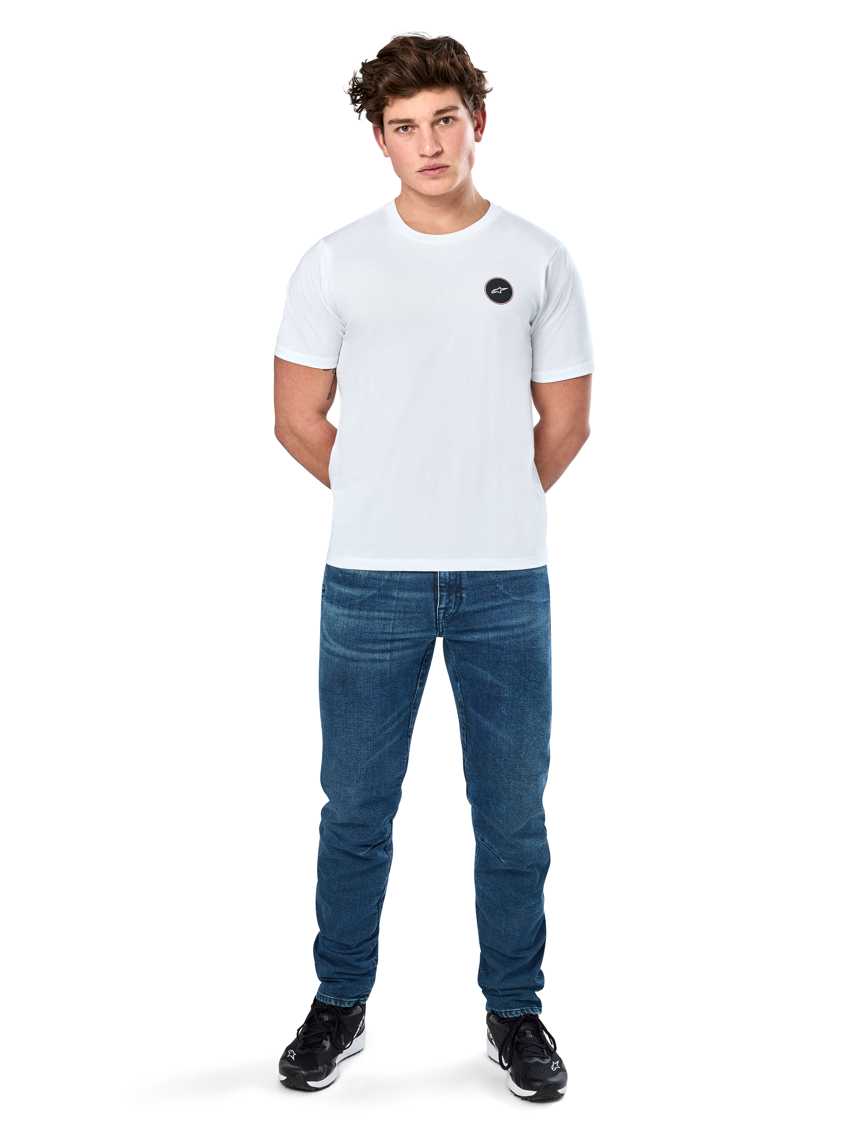 Alpinestars Dot Carbon Csf Tee, camiseta de manga corta, blanca, logotipo circular Astars con efecto de fibra de carbono, ropa casual de estilo automovilismo, usada por un modelo masculino con pantalones vaqueros azules y zapatillas negras