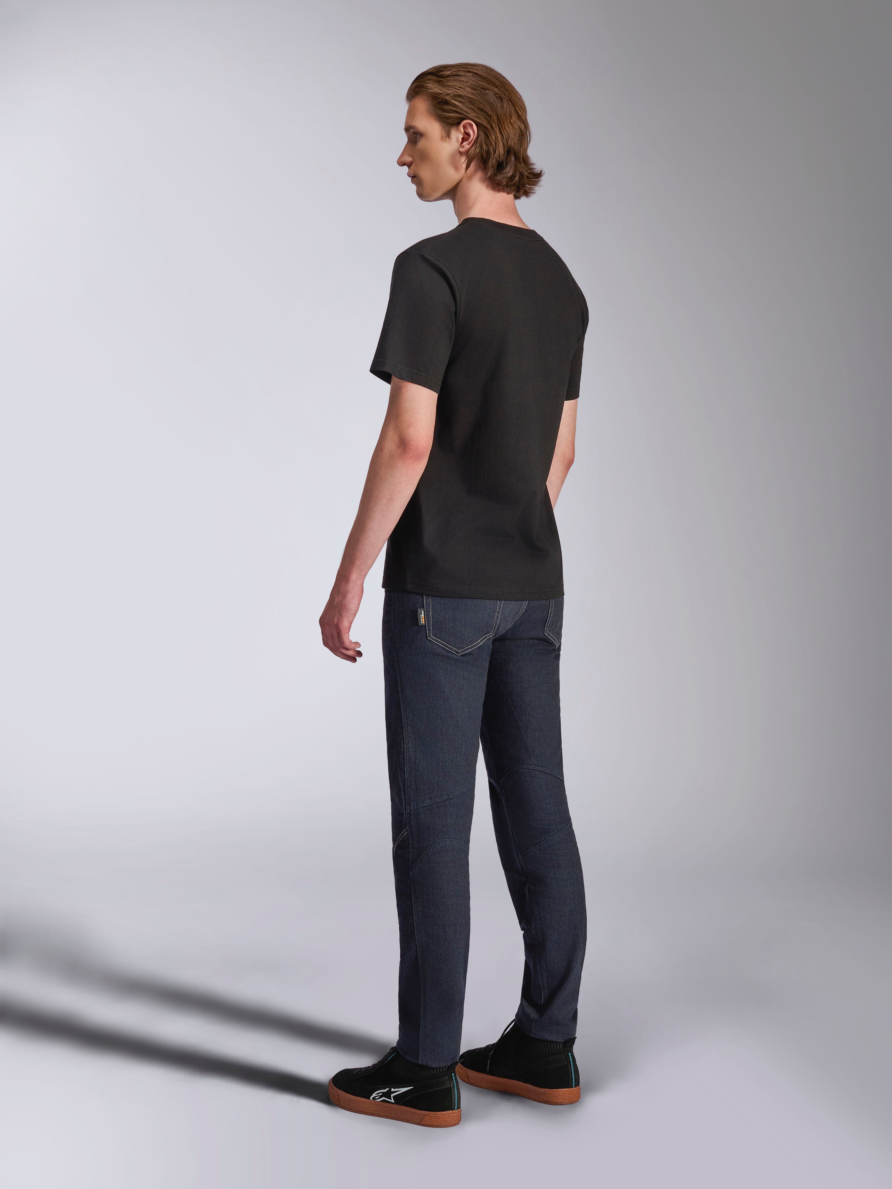 Alpinestars Luxo CSF Tee, camiseta de manga corta, negra, vista posterior en modelo combinada con vaqueros azul oscuro y zapatillas negras, ropa urbana casual inspirada en el automovilismo