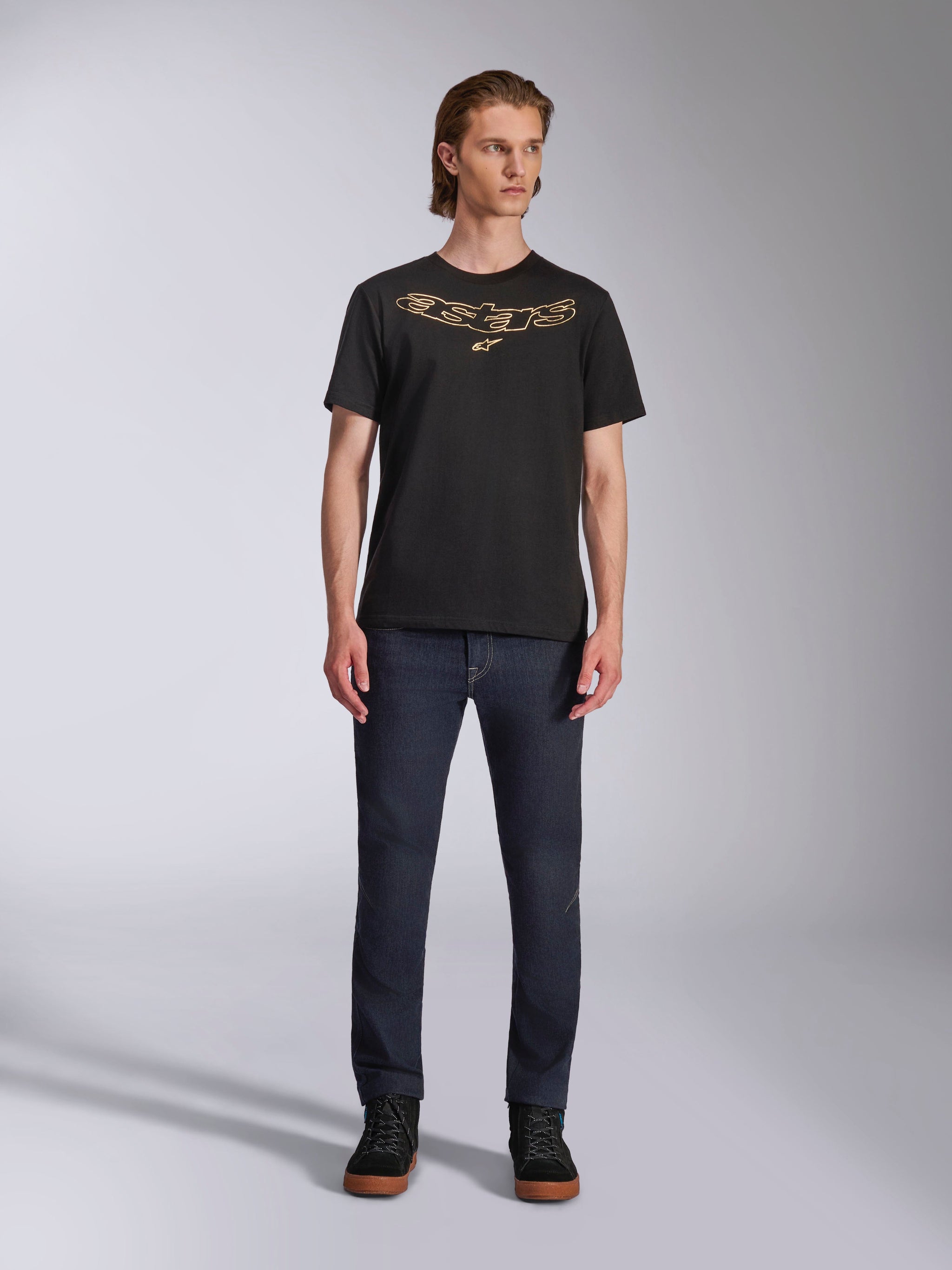 Alpinestars Luxo CSF Tee, camiseta de manga corta, negra, logotipo de Astars contorneado en color canela en el pecho, usada por un modelo con vaqueros oscuros y zapatillas altas negras, estilo casual streetwear de motociclismo
