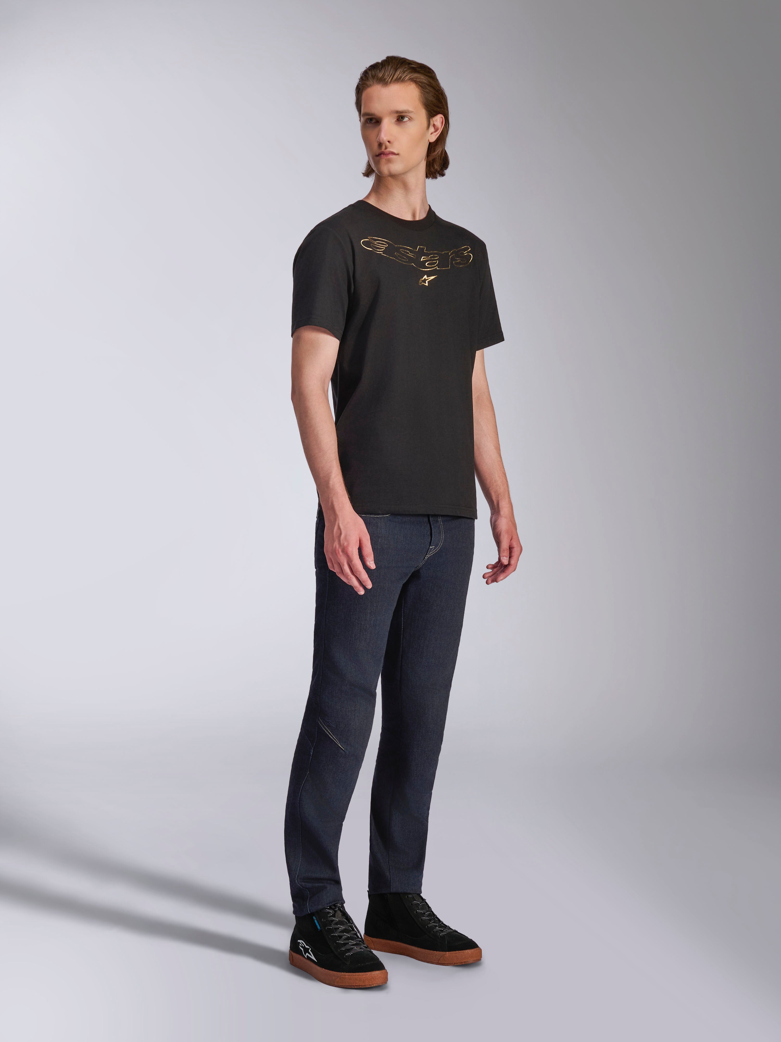 Camiseta Alpinestars Luxo CSF, camiseta de manga corta, negra, logotipo de Astars perfilado en dorado en el pecho, usada por un modelo con vaqueros oscuros y zapatillas negras, ropa de calle casual de automovilismo