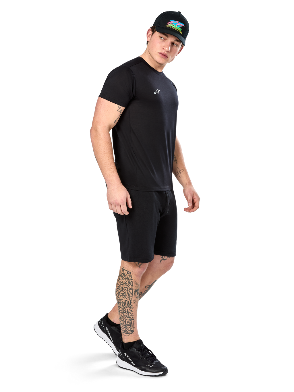 Hombre con tatuajes vistiendo la camiseta Alpinestars Point Performance, camiseta técnica negra de manga corta con un pequeño logo blanco, combinada con pantalones cortos negros, una gorra negra con gráfico colorido y zapatillas blancas y negras, estilo de vida deportivo casual