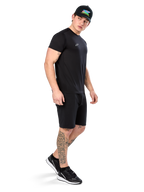 Hombre con tatuajes vistiendo la camiseta Alpinestars Point Performance, camiseta técnica negra de manga corta con un pequeño logo blanco, combinada con pantalones cortos negros, una gorra negra con gráfico colorido y zapatillas blancas y negras, estilo de vida deportivo casual