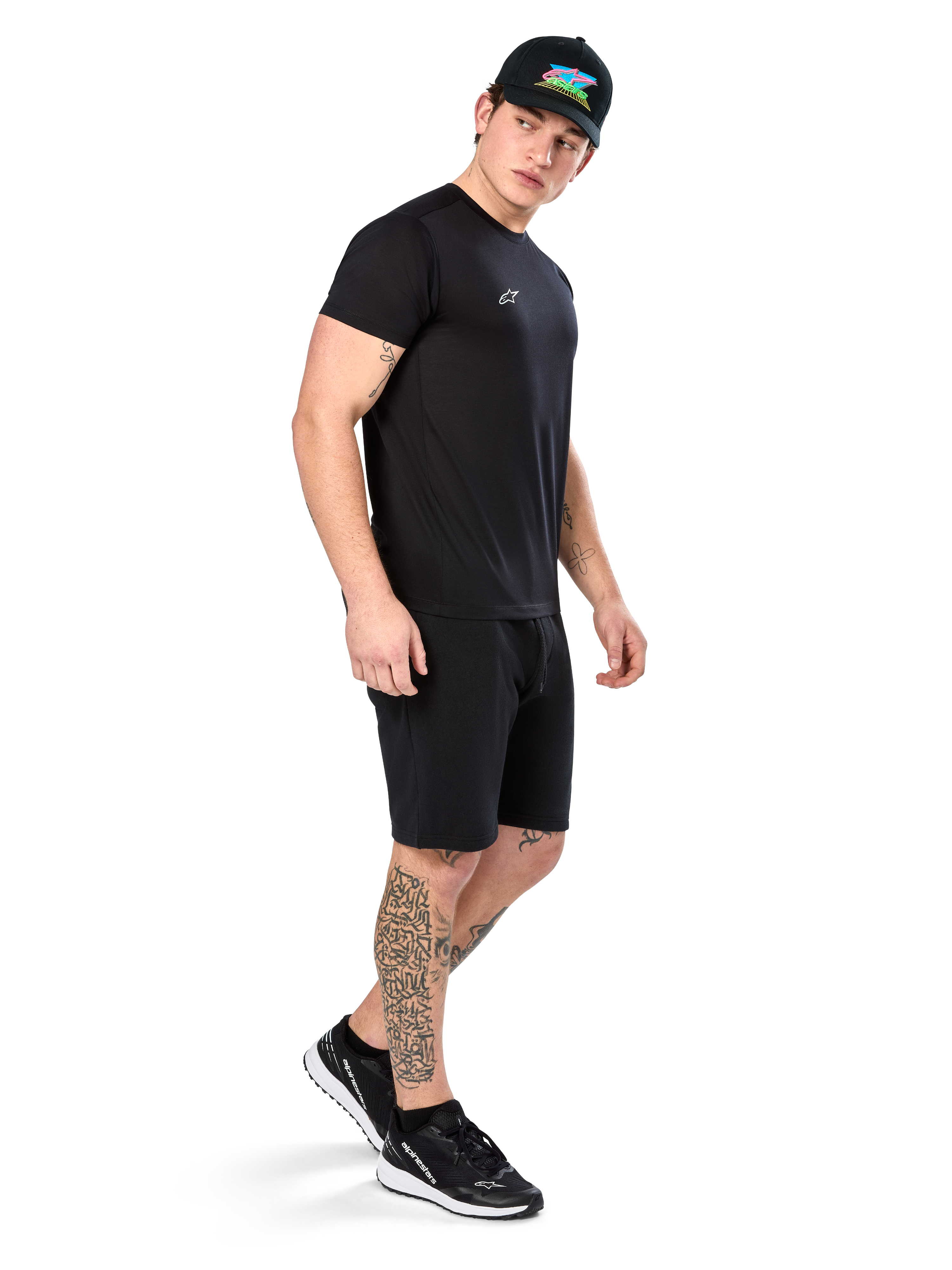 Hombre con tatuajes vistiendo la camiseta Alpinestars Point Performance, camiseta técnica negra de manga corta con un pequeño logo blanco, combinada con pantalones cortos negros, una gorra negra con gráfico colorido y zapatillas blancas y negras, estilo de vida deportivo casual
