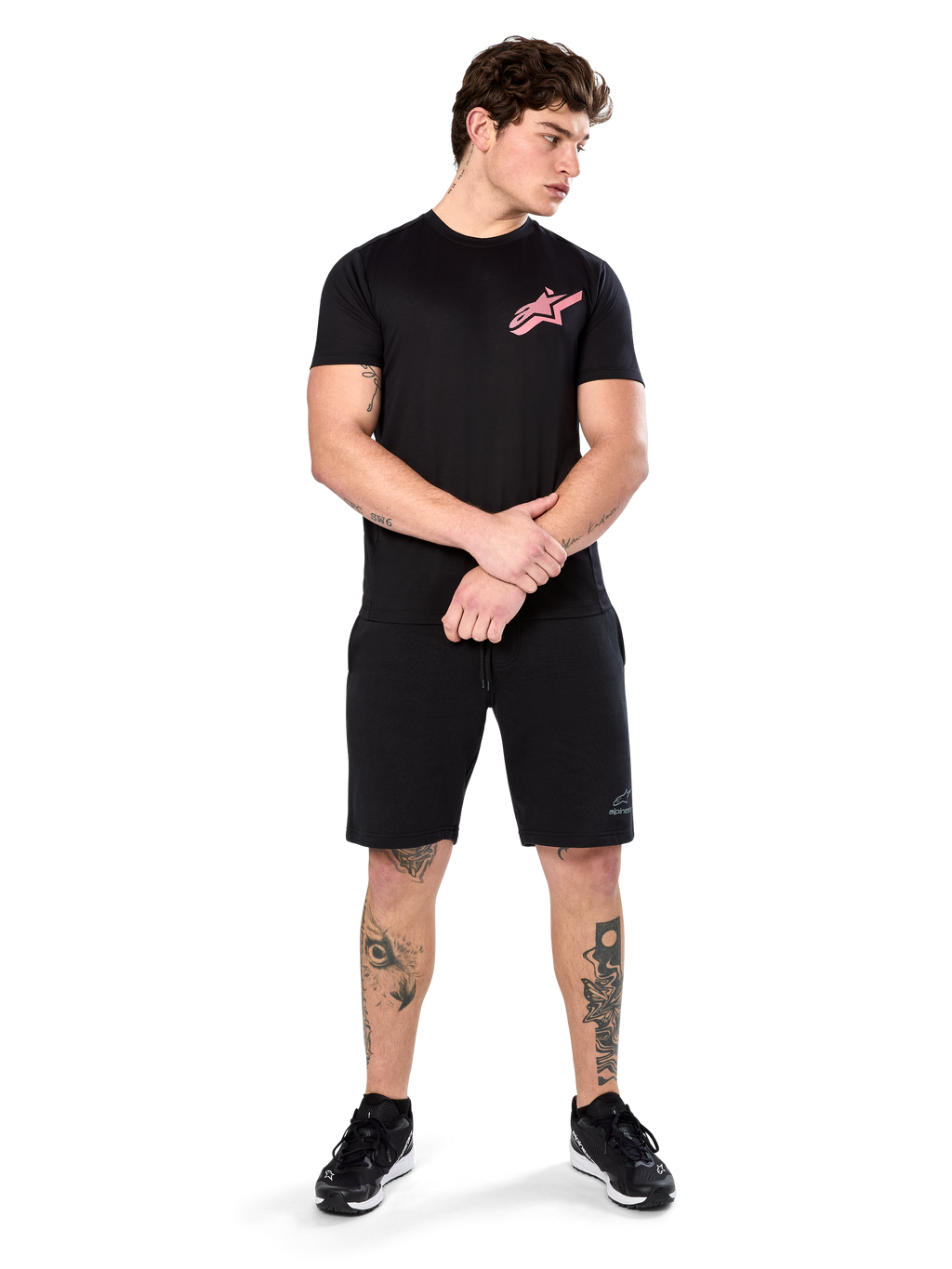 Modelo vistiendo la Alpinestars Attrition Performance Tee, camiseta de rendimiento de manga corta, negra con el logotipo de Astars en rosa en el pecho, combinada con pantalones cortos negros y zapatillas, ropa deportiva casual inspirada en el automovilismo