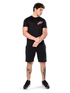 Modelo vistiendo la Alpinestars Attrition Performance Tee, camiseta de rendimiento de manga corta, negra con el logotipo de Astars en rosa en el pecho, combinada con pantalones cortos negros y zapatillas, ropa deportiva casual inspirada en el automovilismo