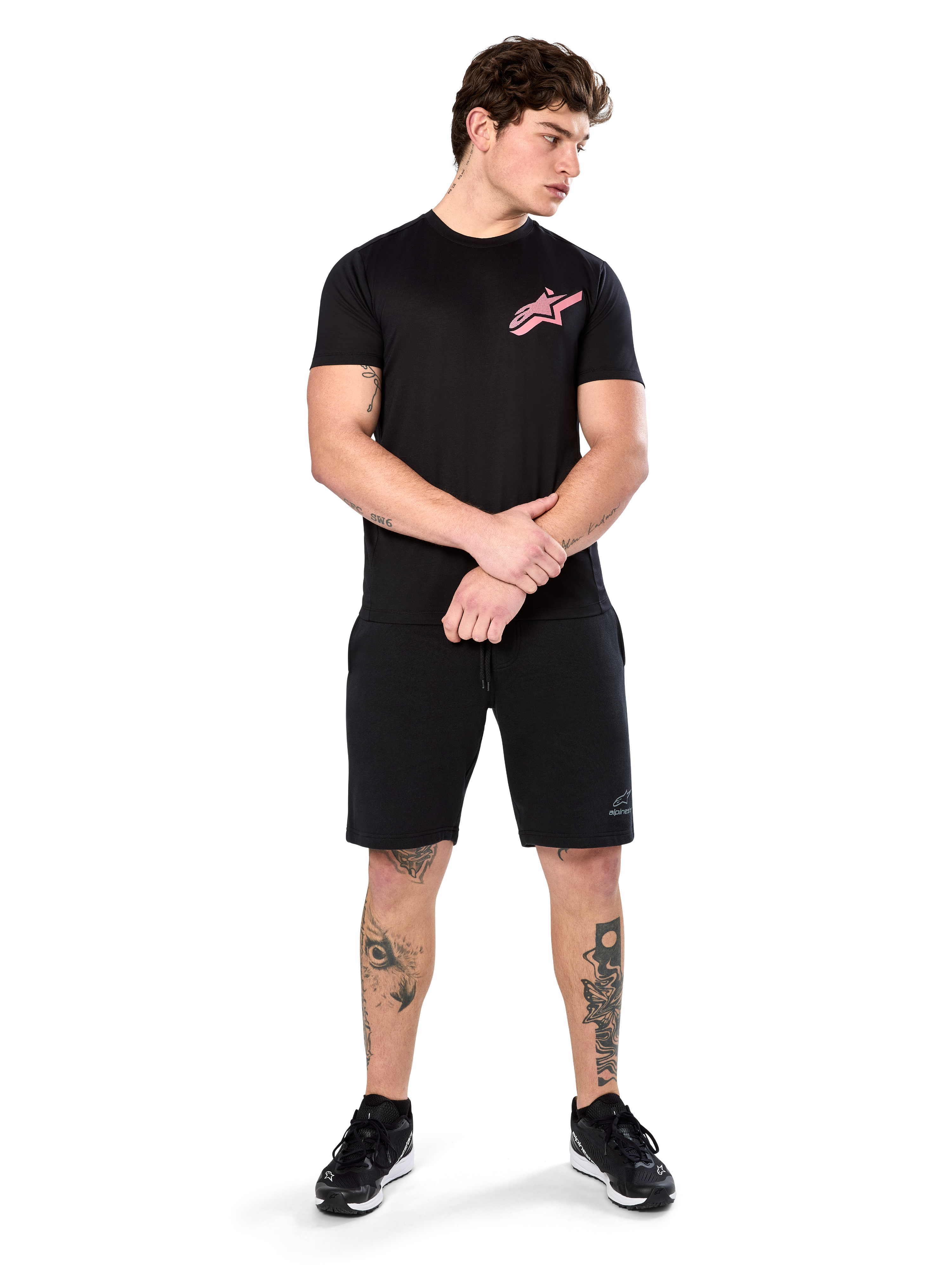 Modelo vistiendo la Alpinestars Attrition Performance Tee, camiseta de rendimiento de manga corta, negra con el logotipo de Astars en rosa en el pecho, combinada con pantalones cortos negros y zapatillas, ropa deportiva casual inspirada en el automovilismo