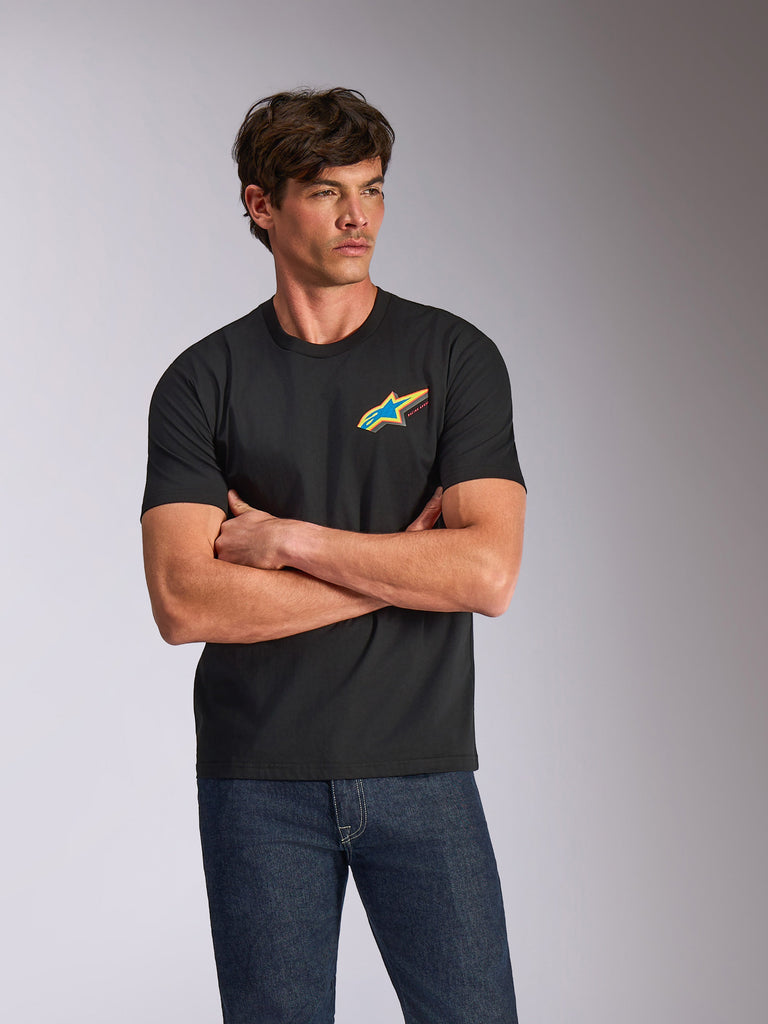 Alpinestars Angler CSF Tee, camiseta de manga corta, negra, con un vibrante logotipo Astars multicolor en el pecho, lucida por un modelo con los brazos cruzados y vaqueros azules, ropa de calle casual inspirada en el automovilismo