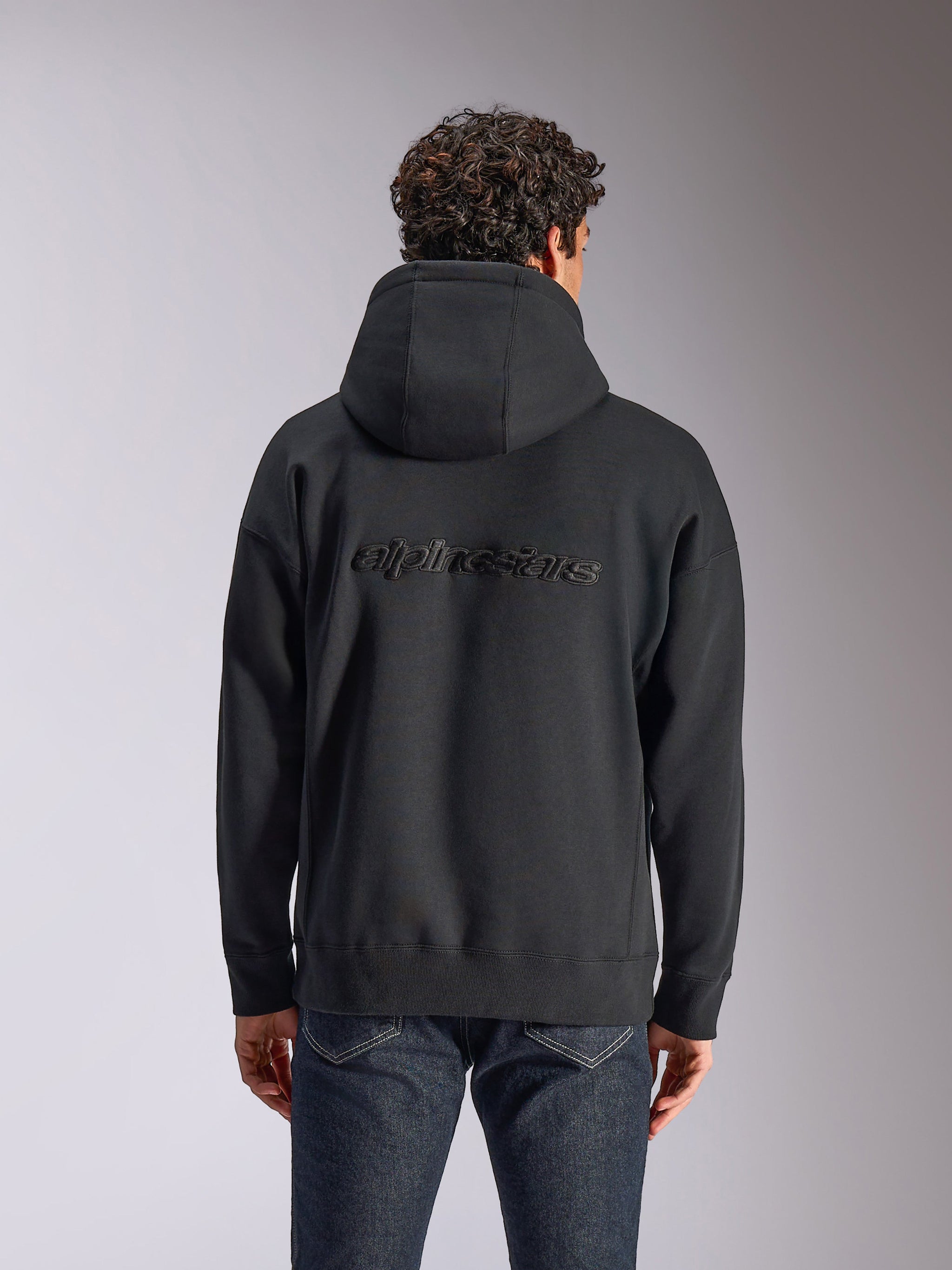 Alpinestars Emergent Oversized Hoodie, vista trasera de una sudadera negra con capucha en un modelo, con el logotipo de Alpinestars bordado en la espalda, ajuste oversized, estilo urbano casual combinado con jeans oscuros