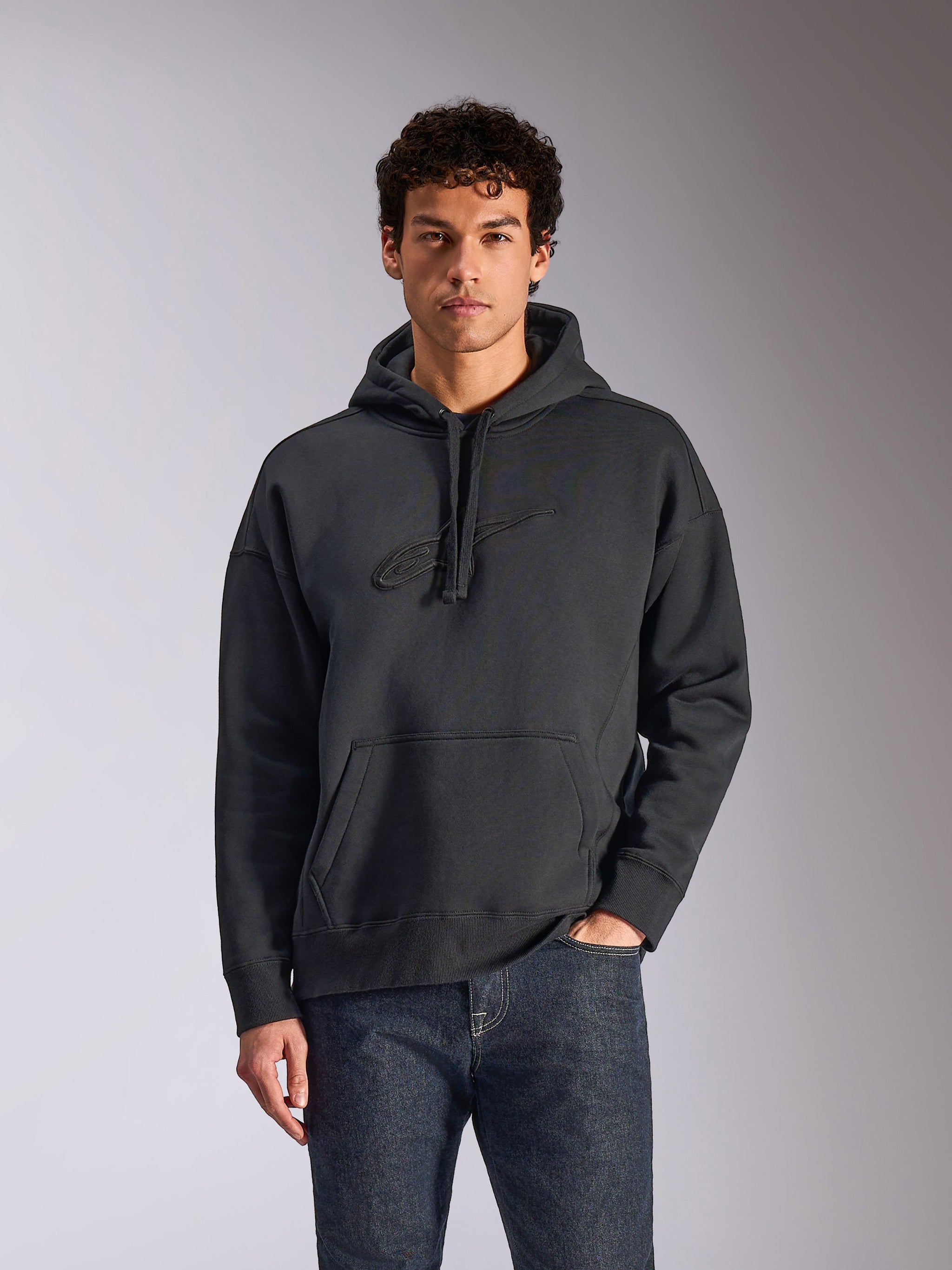 Sudadera Emergent Oversized de Alpinestars, negra, modelo masculino con sudadera de forro polar de corte holgado, logotipo Astars bordado en 3D a tono, bolsillo canguro, capucha con cordón, ropa urbana informal inspirada en el motociclismo