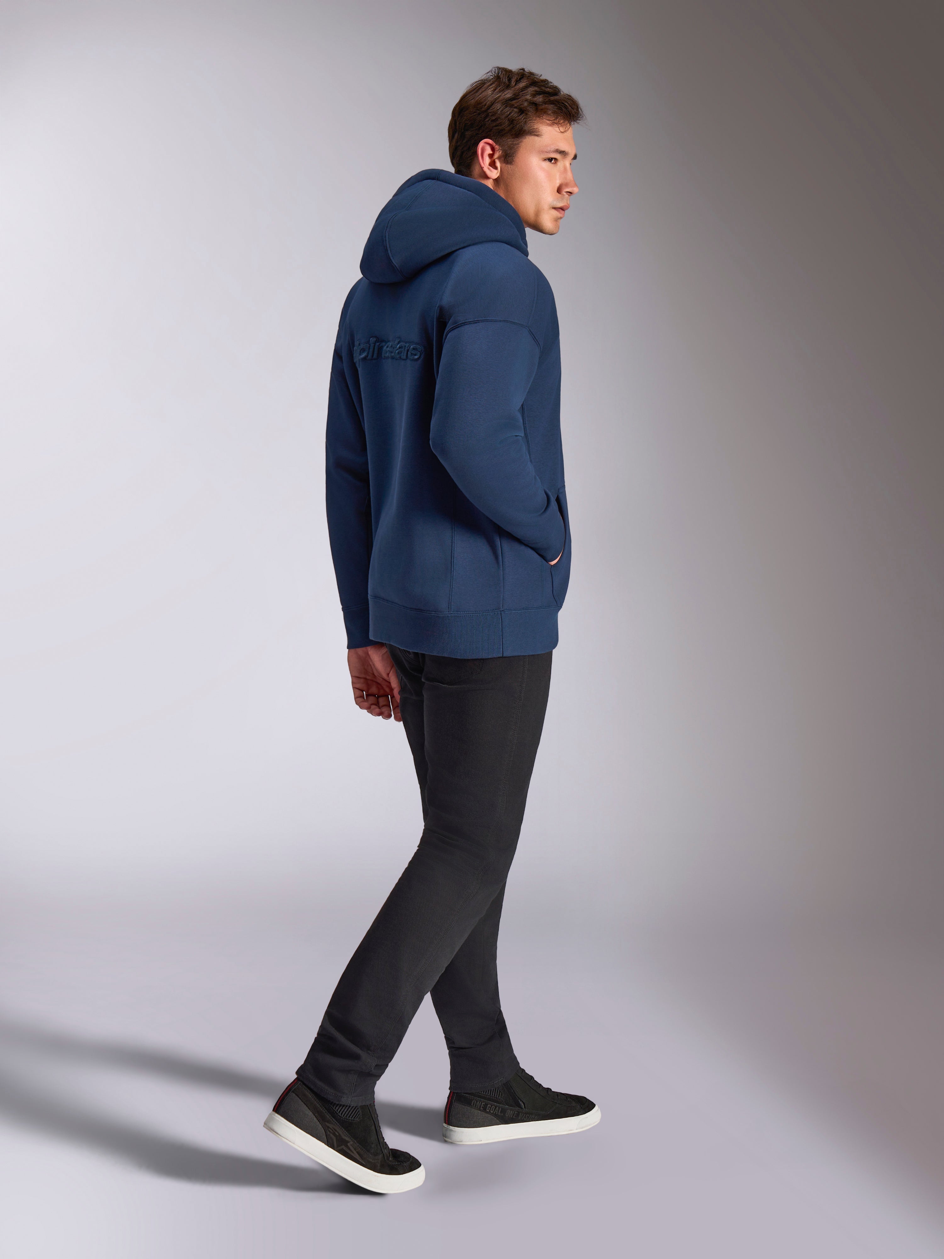Alpinestars Emergent Oversized Hoodie in Navy Blue, back view showing 3D embroidered Alpinestars word logo, oversized fleece fit with drawstring hood, worn with black pants, casual motorsport-inspired streetwear