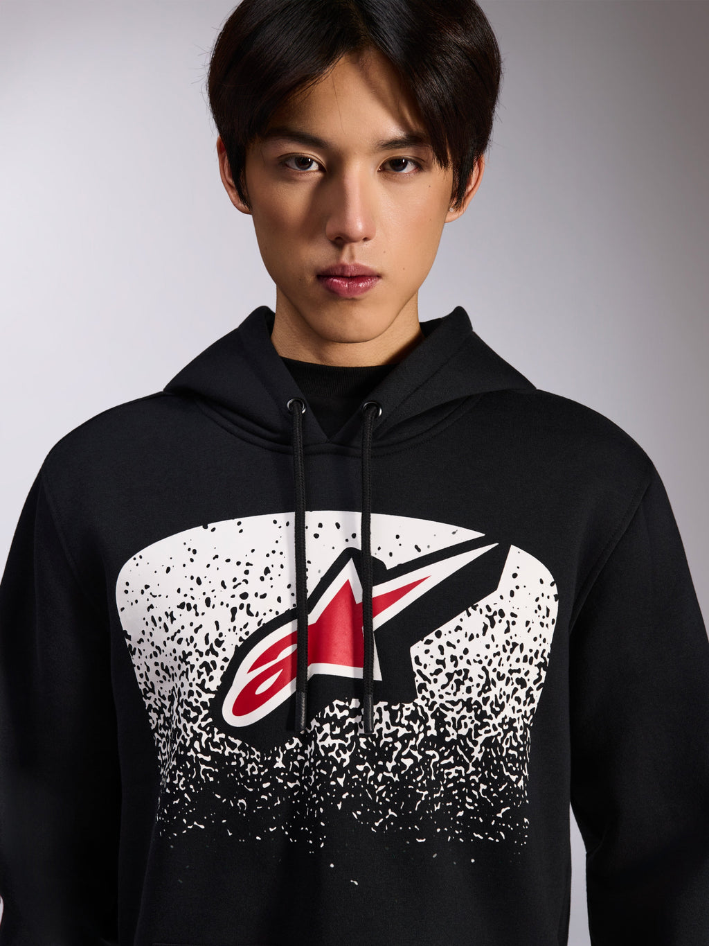 Sudadera con capucha Alpinestars Obsessed, sudadera negra de felpa, con un logotipo Astars rojo y blanco dentro de un gráfico degradado MX punteado en blanco en el pecho, usada por un modelo, estilo casual motorsport con capucha ajustable con cordón
