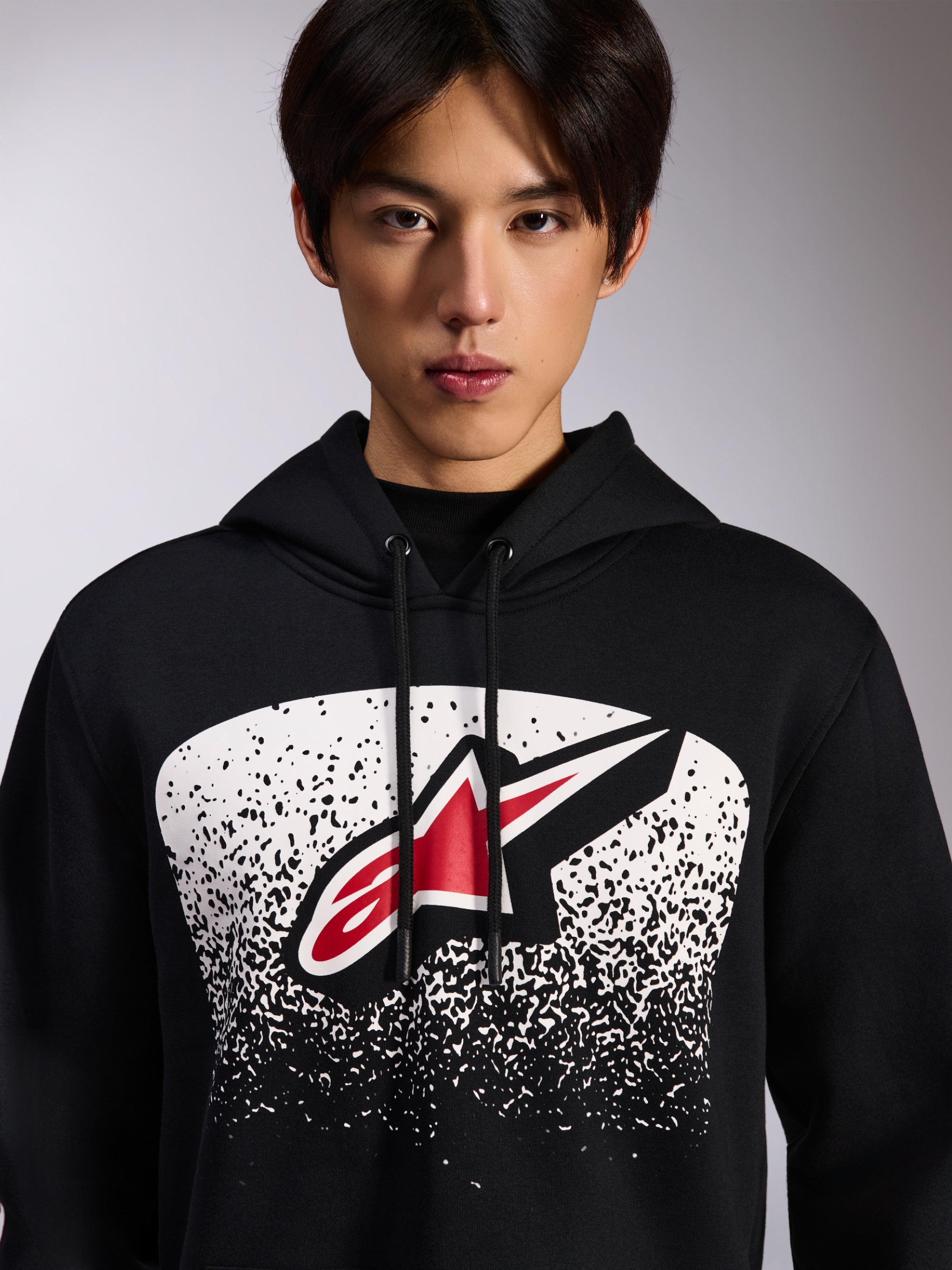 Sudadera con capucha Alpinestars Obsessed, sudadera negra de felpa, con un logotipo Astars rojo y blanco dentro de un gráfico degradado MX punteado en blanco en el pecho, usada por un modelo, estilo casual motorsport con capucha ajustable con cordón