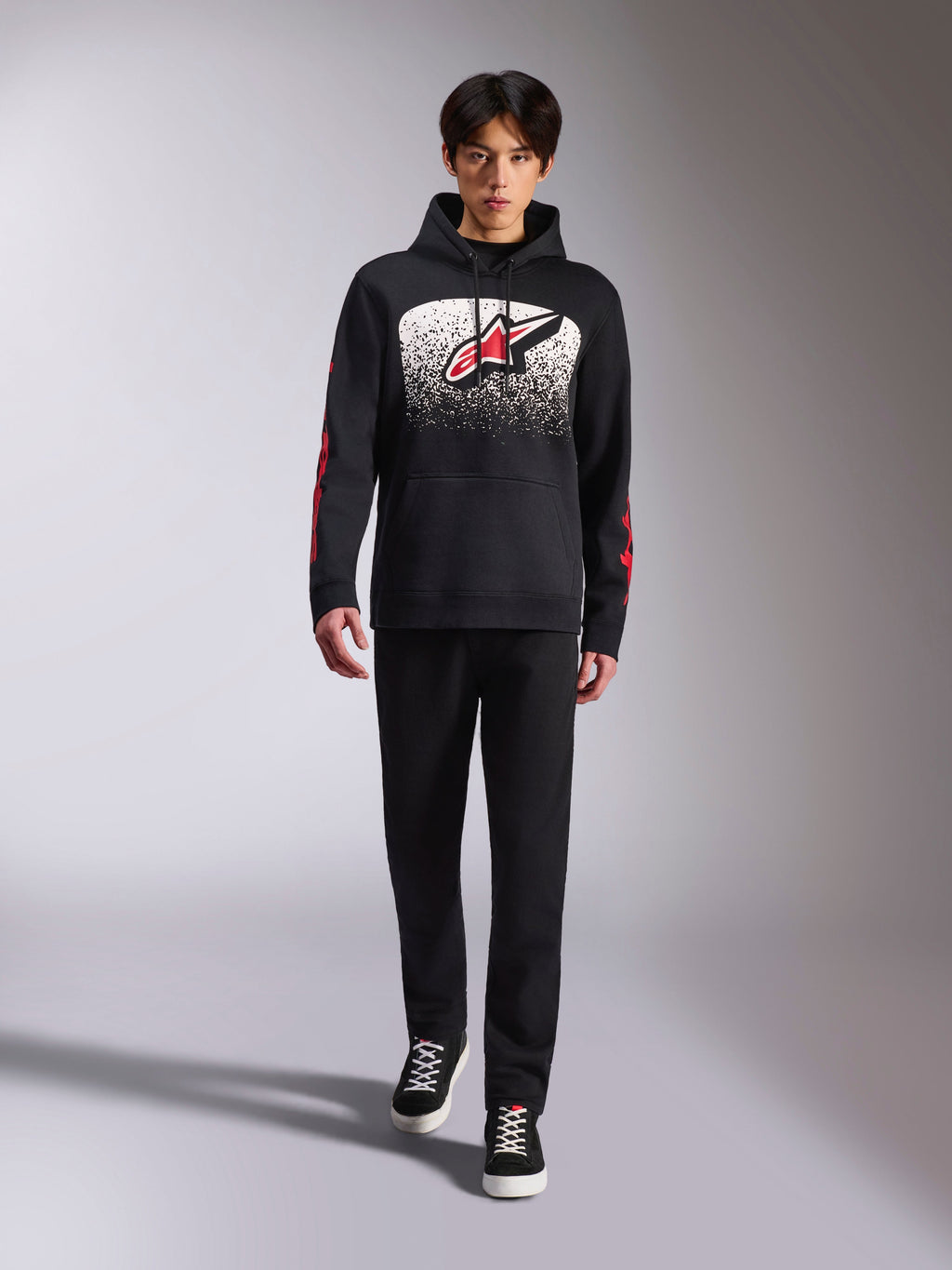Sudadera con capucha Alpinestars Obsessed, sudadera negra tipo pullover, logotipo grande de Astars en blanco y rojo con degradado MX, logotipos rojos en las mangas, modelo con conjunto casual totalmente negro, ropa lifestyle inspirada en el motociclismo