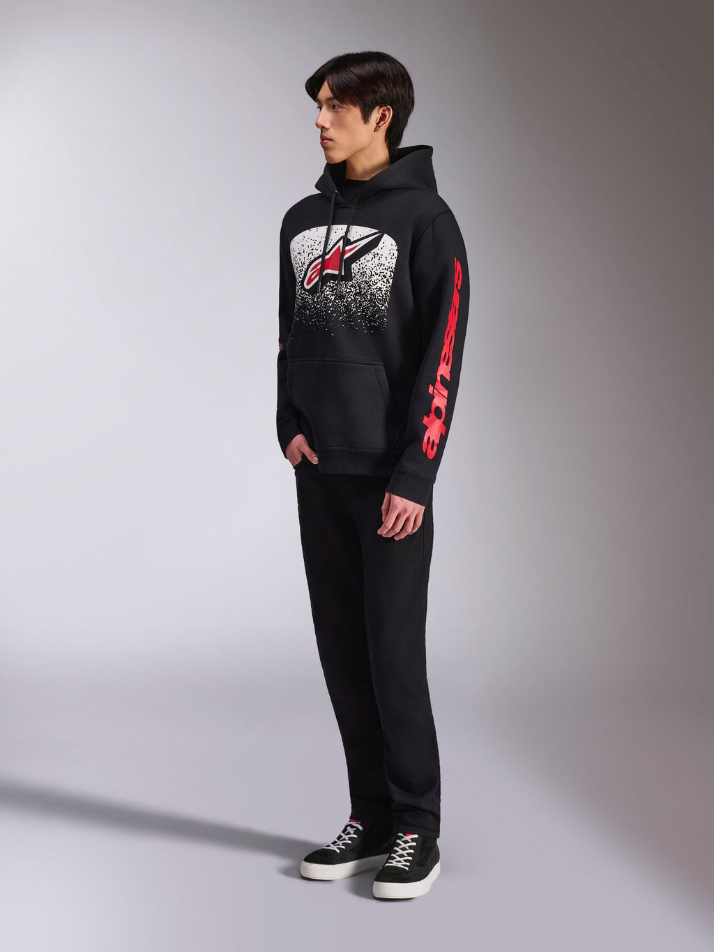 Alpinestars Obsessed Hoodie, sudadera de forro polar negro con un gran gráfico moteado en blanco en el pecho, logotipo Astars en rojo y marca roja en la manga, usada por un modelo con pantalones negros y zapatillas, estilo casual motorsport