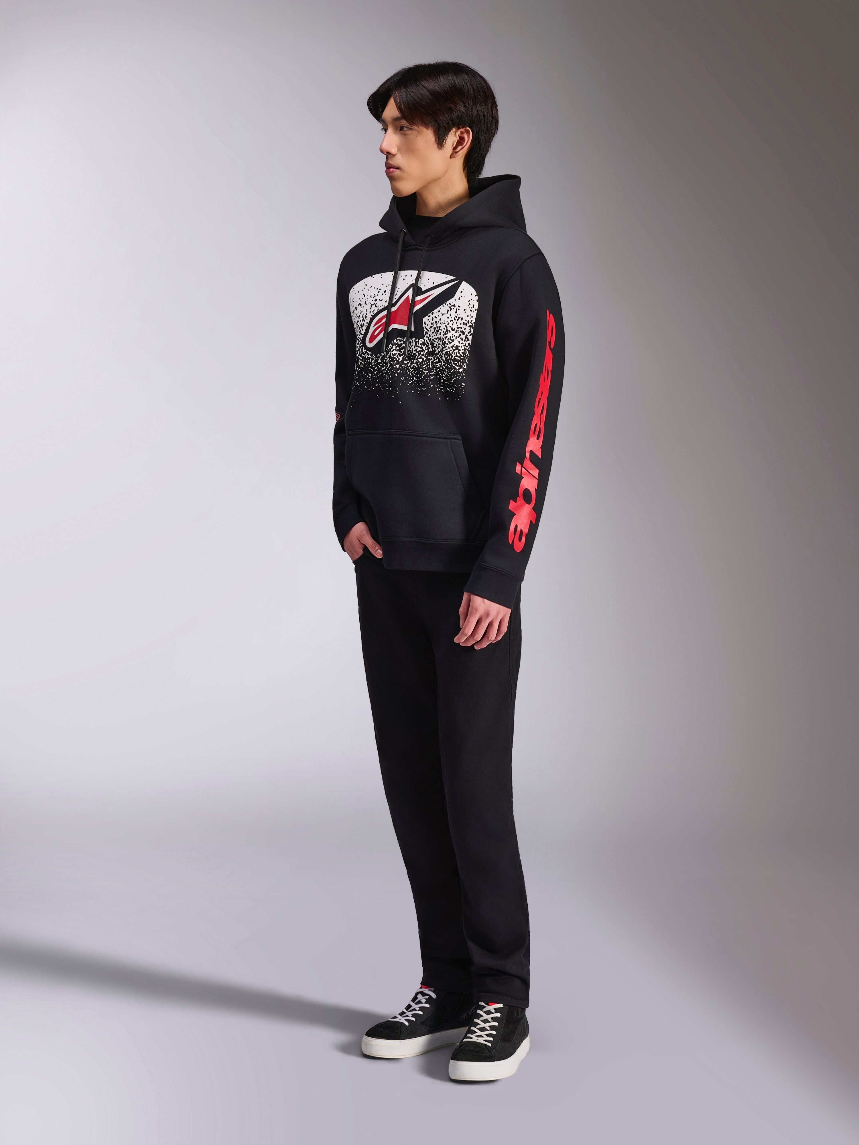 Alpinestars Obsessed Hoodie, sudadera de forro polar negro con un gran gráfico moteado en blanco en el pecho, logotipo Astars en rojo y marca roja en la manga, usada por un modelo con pantalones negros y zapatillas, estilo casual motorsport