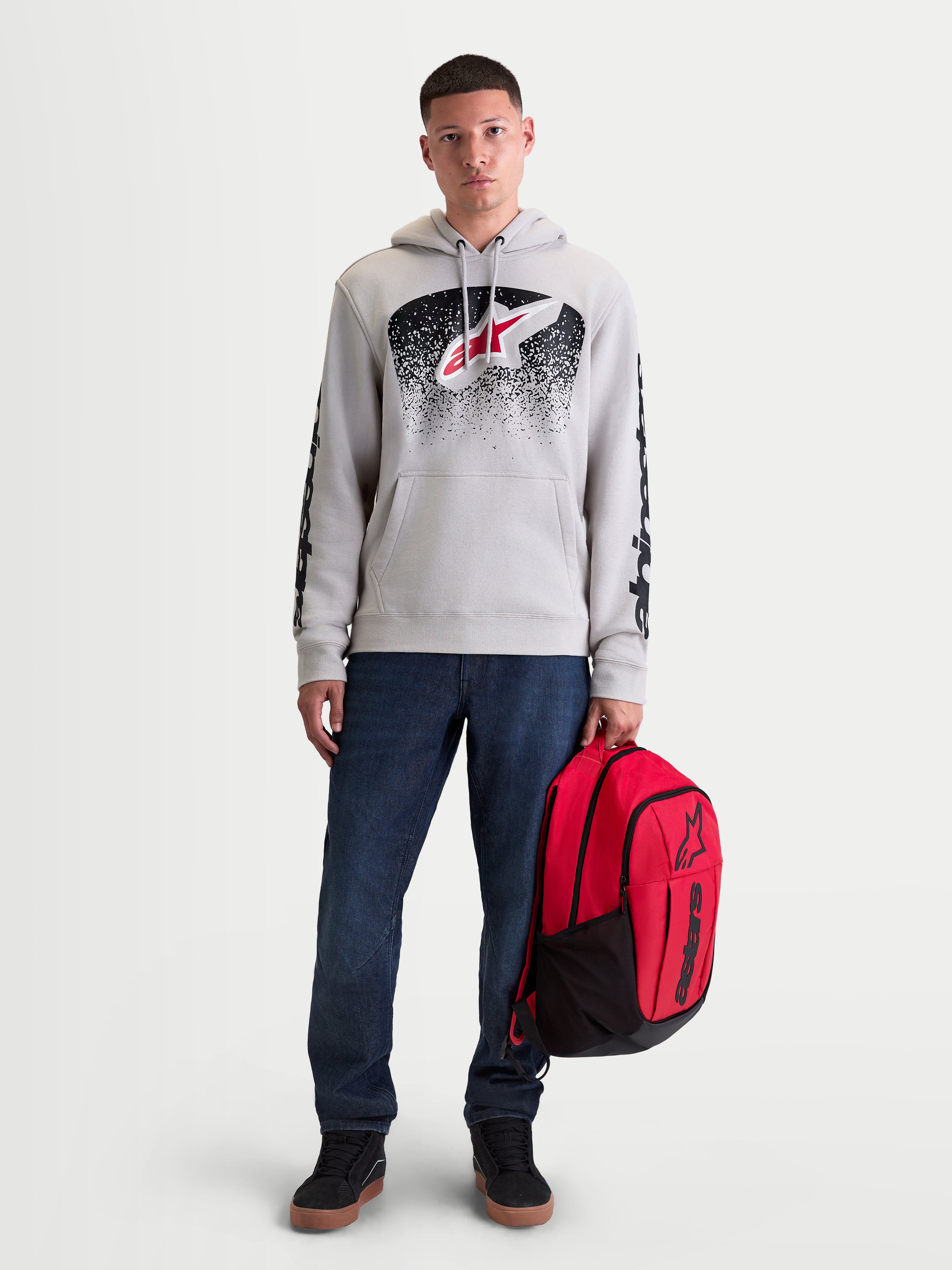 Alpinestars Sudadera con capucha Obsessed, sudadera deportiva para hombre, gris cemento con gráficos degradados en rojo y negro inspirados en el motocross, logotipos en las mangas, mostrada en un modelo con mochila roja