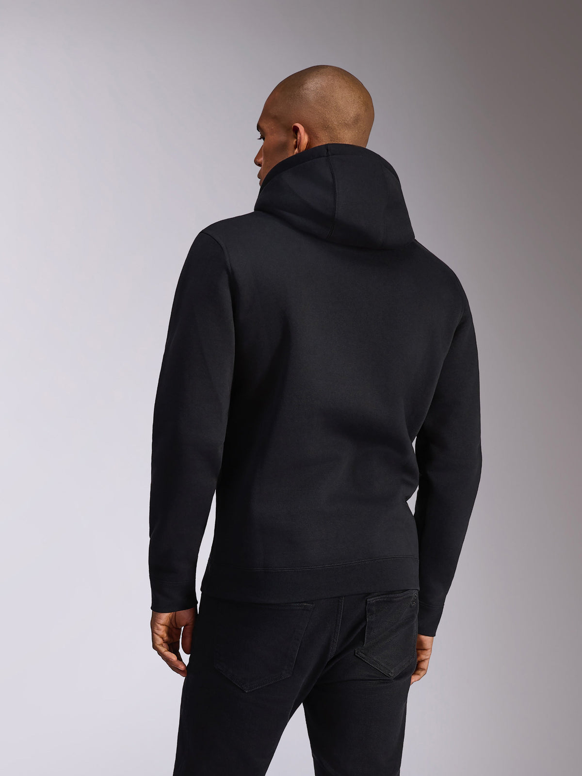 Sudadera Alpinestars Ellipsoid, negra, vista trasera de una persona que lleva una sudadera de felpa con capucha, mangas largas con puños y dobladillo de canalé, combinada con vaqueros negros, ropa deportiva casual inspirada en el motor
