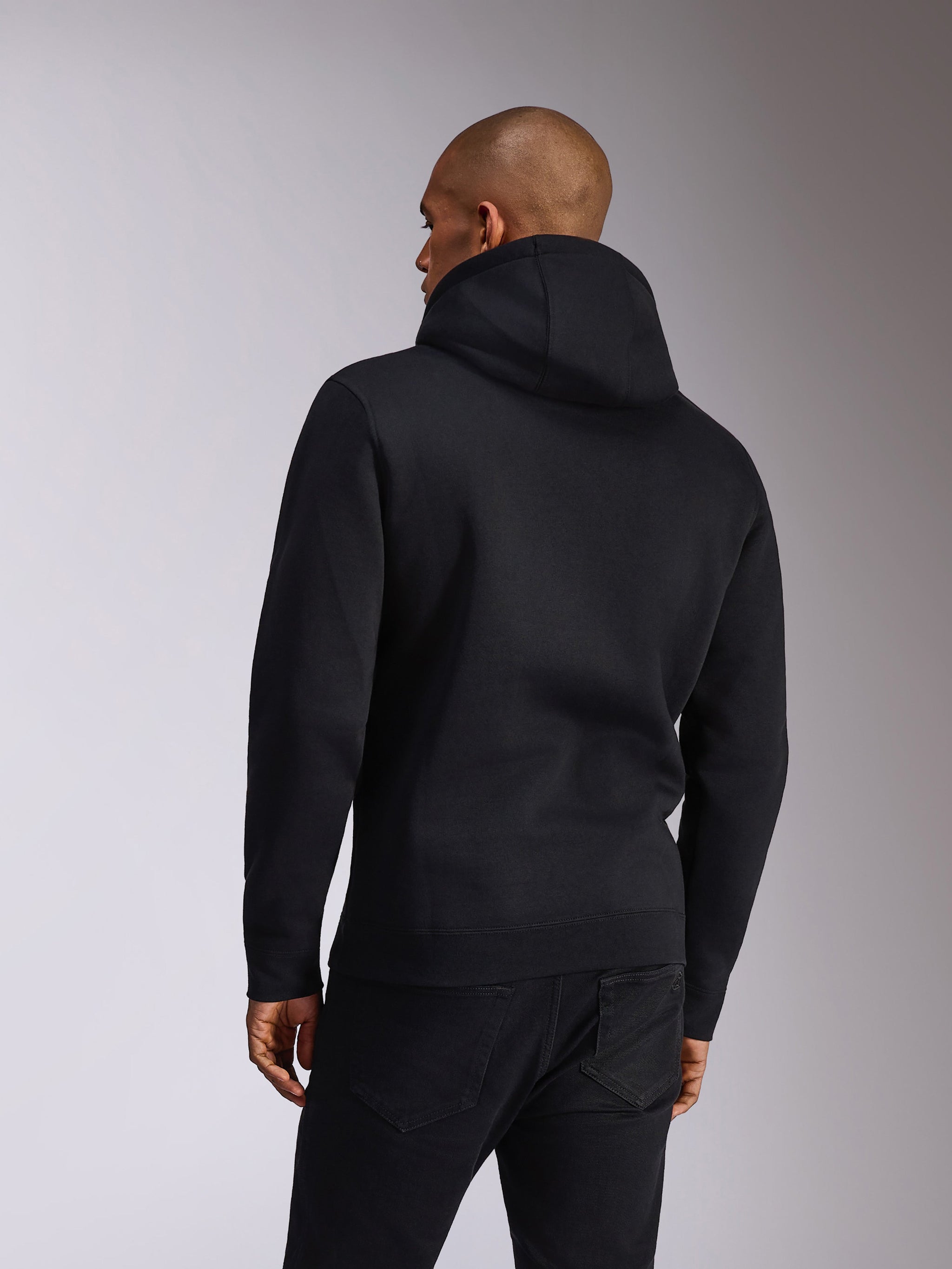 Sudadera Alpinestars Ellipsoid, negra, vista trasera de una persona que lleva una sudadera de felpa con capucha, mangas largas con puños y dobladillo de canalé, combinada con vaqueros negros, ropa deportiva casual inspirada en el motor