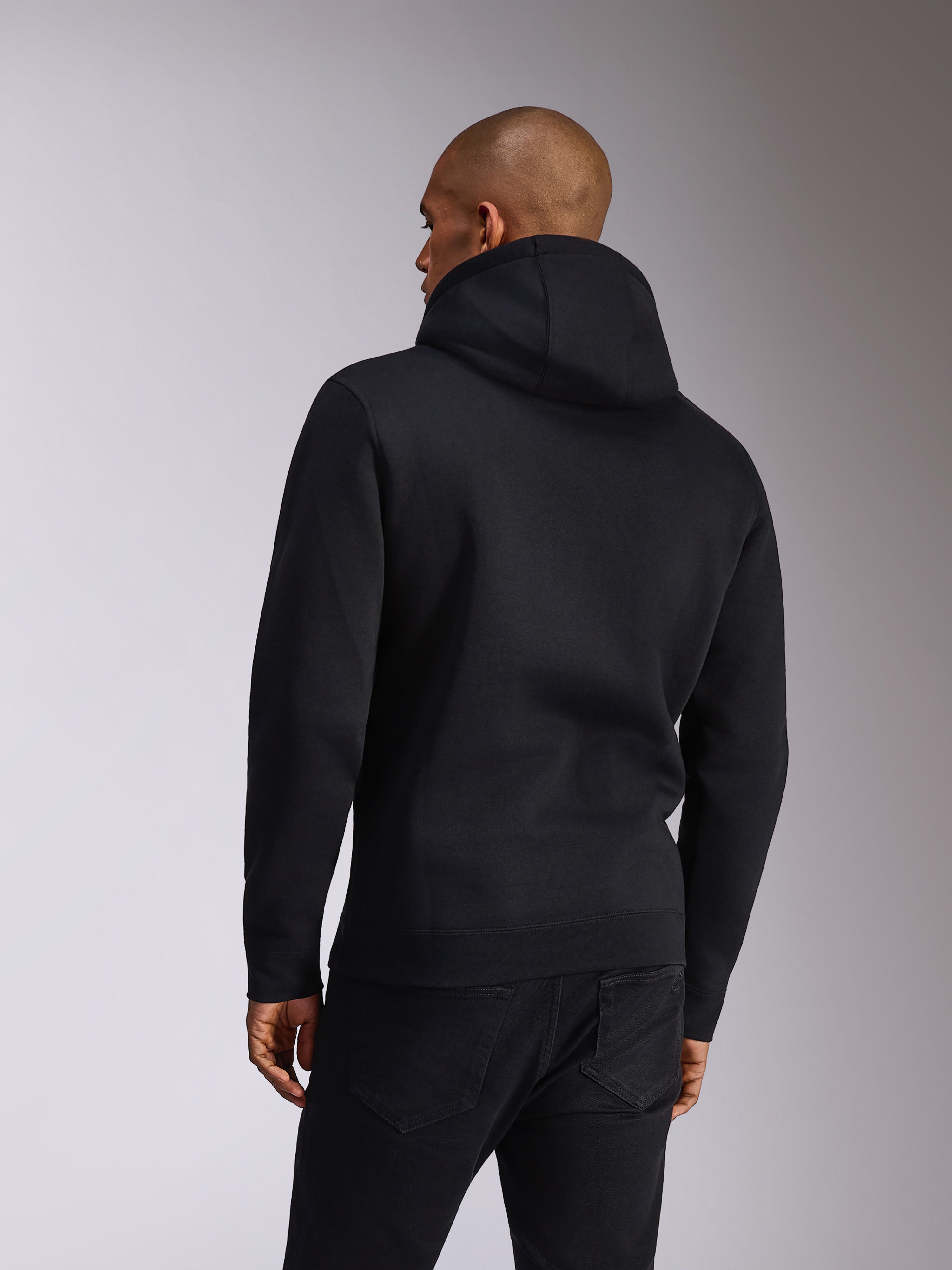 Sudadera Alpinestars Ellipsoid, negra, vista trasera de una persona que lleva una sudadera de felpa con capucha, mangas largas con puños y dobladillo de canalé, combinada con vaqueros negros, ropa deportiva casual inspirada en el motor