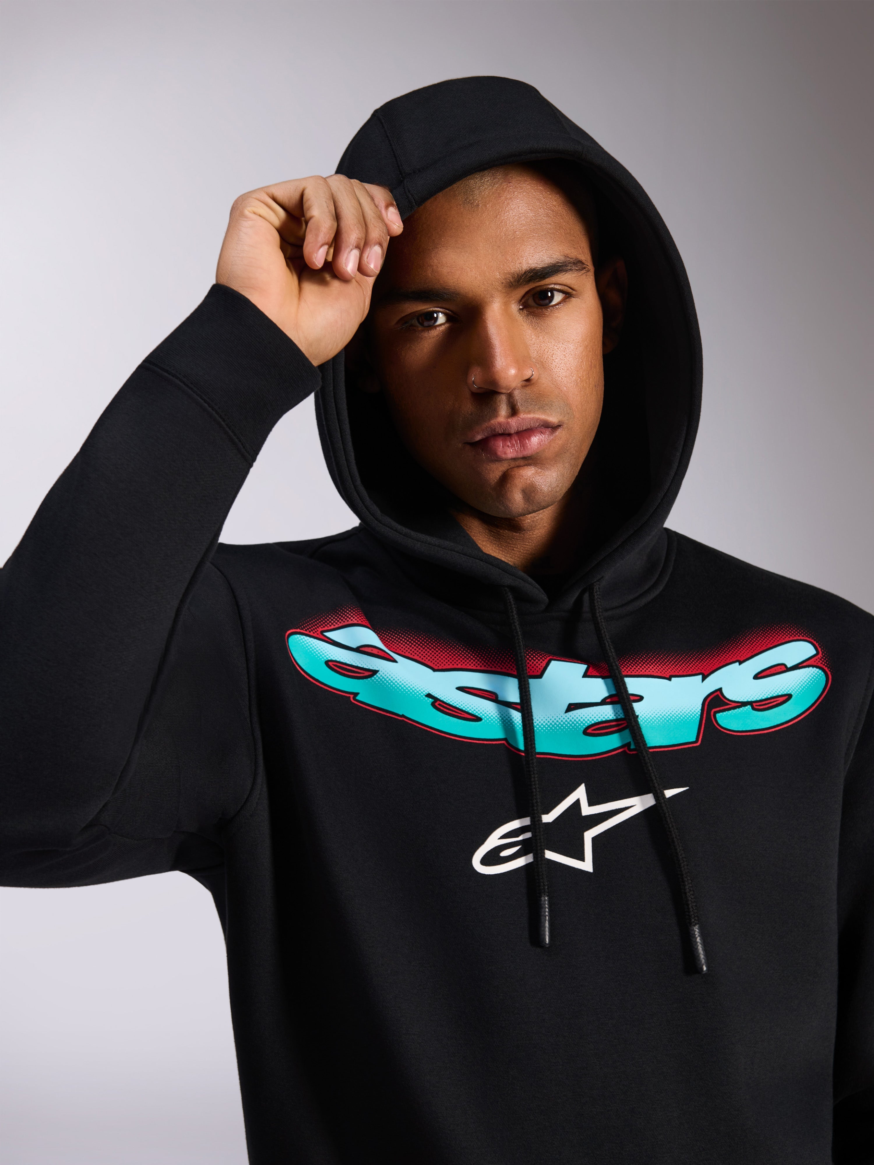 Alpinestars Ellipsoid Hoodie, sudadera negra con capucha, con la palabra Astars estilizada en degradado de color verde azulado y rojo y logotipo blanco en el pecho, lucida por un modelo con la capucha puesta, ropa de calle informal inspirada en el automovilismo