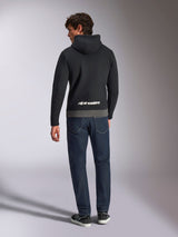 Sudadera con capucha Alpinestars Parallel, vista trasera de un hombre con un forro polar de tipo pullover en bloques de color negro y gunmetal, logotipo de Alpinestars en blanco en la parte inferior de la espalda, combinado con vaqueros azul oscuro, ropa urbana casual inspirada en el motociclismo