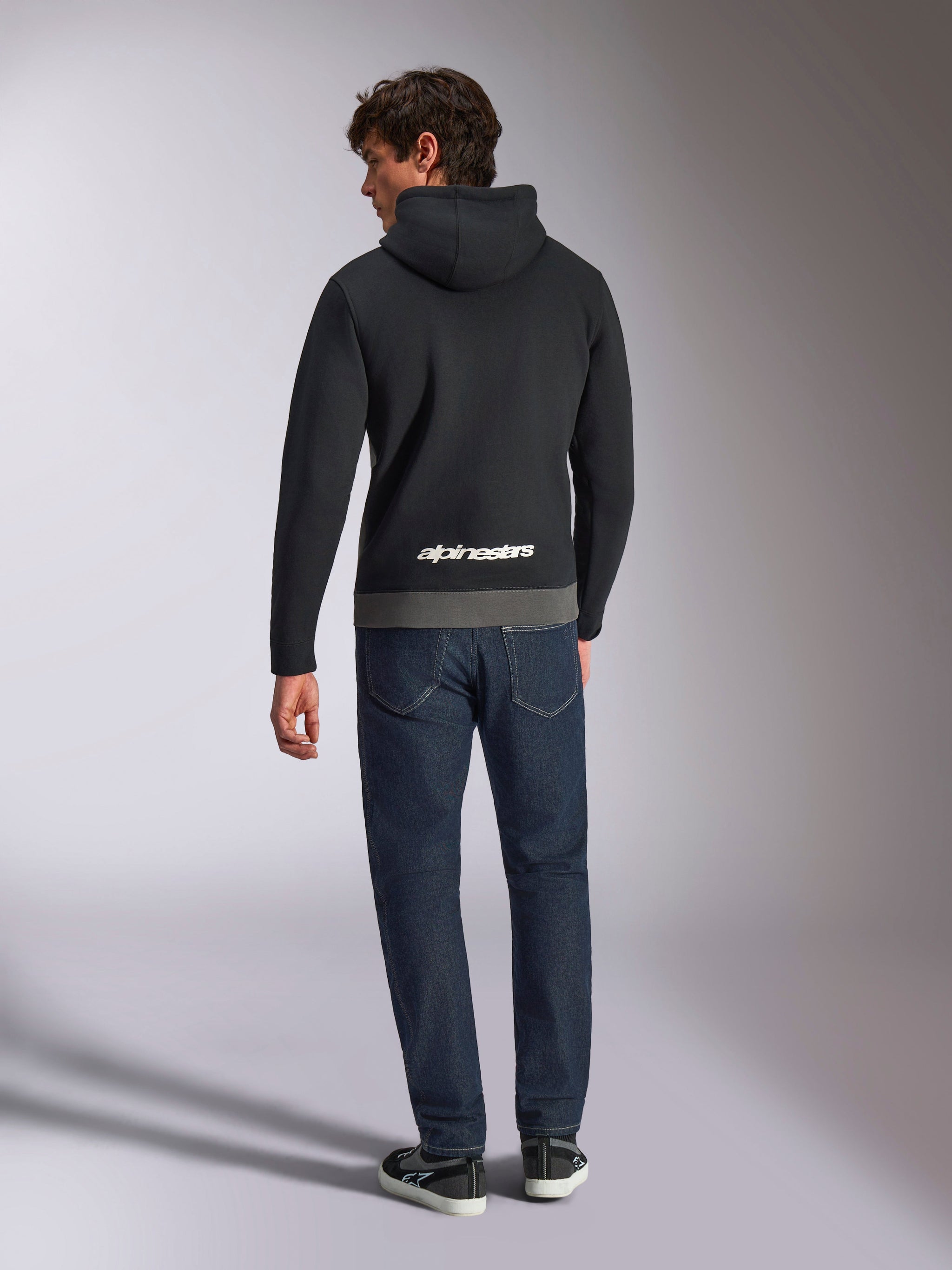 Sudadera con capucha Alpinestars Parallel, vista trasera de un hombre con un forro polar de tipo pullover en bloques de color negro y gunmetal, logotipo de Alpinestars en blanco en la parte inferior de la espalda, combinado con vaqueros azul oscuro, ropa urbana casual inspirada en el motociclismo