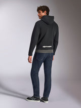 Sudadera Parallel de Alpinestars, vista posterior del modelo con el jersey en negro y gris plomo, con el logotipo blanco de Alpinestars en la parte inferior de la espalda, combinado con vaqueros azul oscuro, estilo urbano casual inspirado en el motor