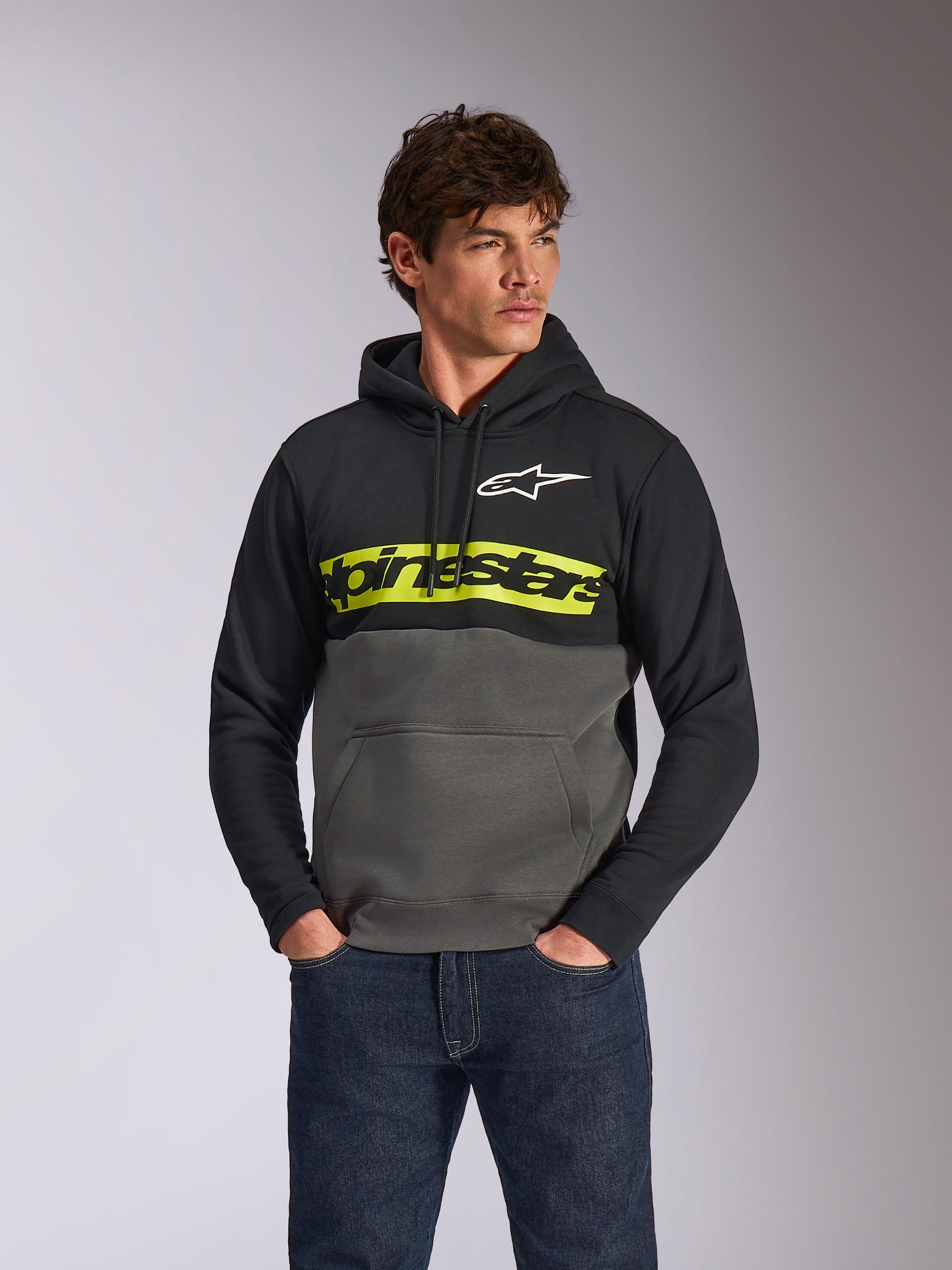 Sudadera con capucha Alpinestars Parallel, sudadera casual con bloques de color, negro y gris plomo con una franja amarilla vibrante en el pecho, logotipo de Alpinestars y logo de estrella blanca, usada por un modelo masculino con vaqueros azul oscuro